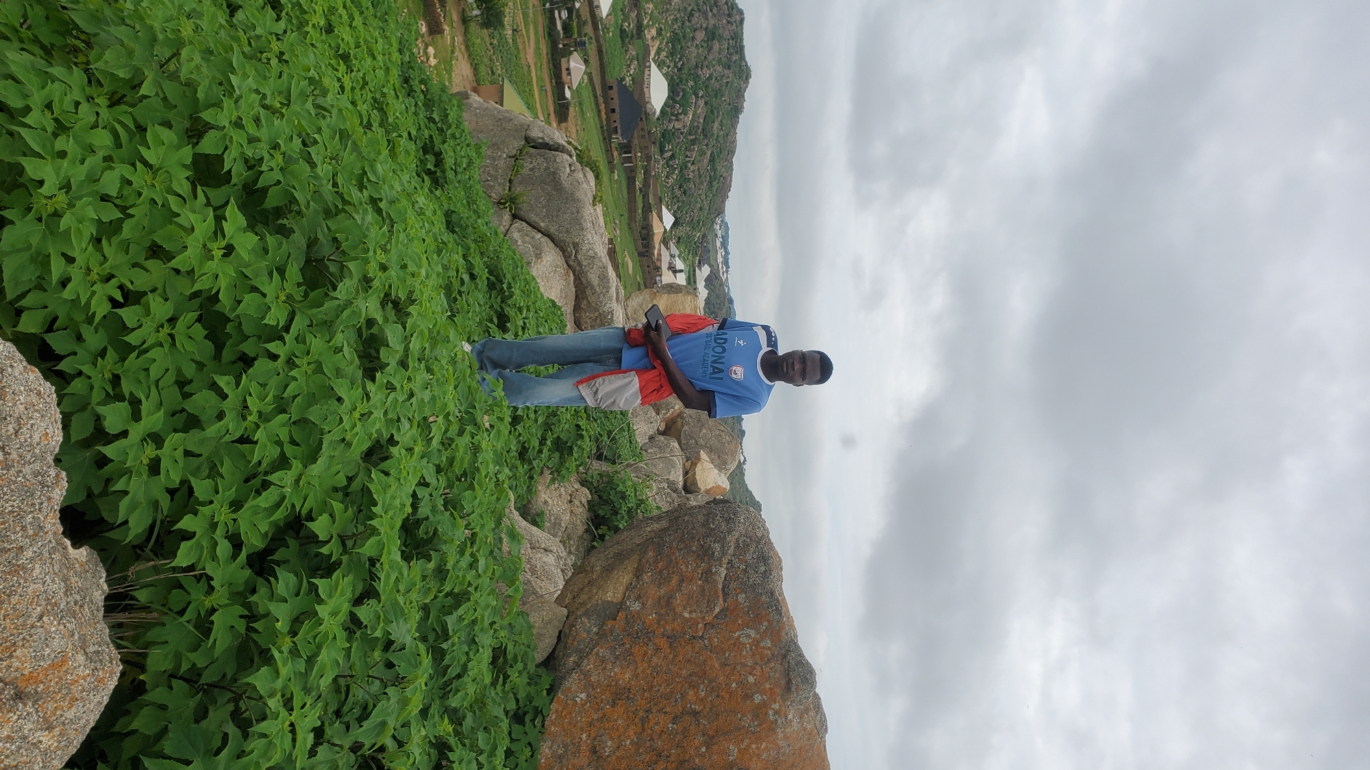 A young Black male stands on a rocky, green hillside overlooking a village. He is wearing a blue t-shirt with the words "ADONAI FOOTBALL ACADEMY" printed on it, blue jeans, and a red and grey jacket tied around his waist. He is holding a smartphone in his right hand. The hillside is covered in lush green vegetation and large rocks, one of which is prominent in the foreground and another to his right. In the background, several houses with distinct roof structures are visible, nestled among rolling hills. The sky is overcast with a dull grey cloud cover. This scene is located in the city of Jos, Nigeria.