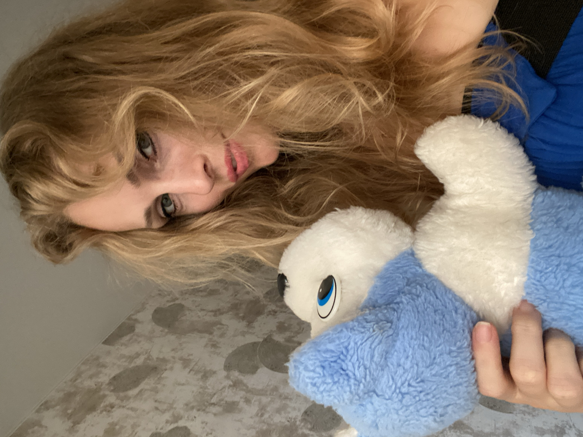 A woman with long, wavy blonde hair and light blue eyes is holding a blue and white stuffed toy in front of her face. She is looking towards the camera with her lips slightly parted. She is wearing a blue garment. The background consists of a textured wall and a portion of a ceiling. The setting is indoors, in a room. There is no visual information in the image that would allow for the determination of a specific location within the city of Lida, Belarus.