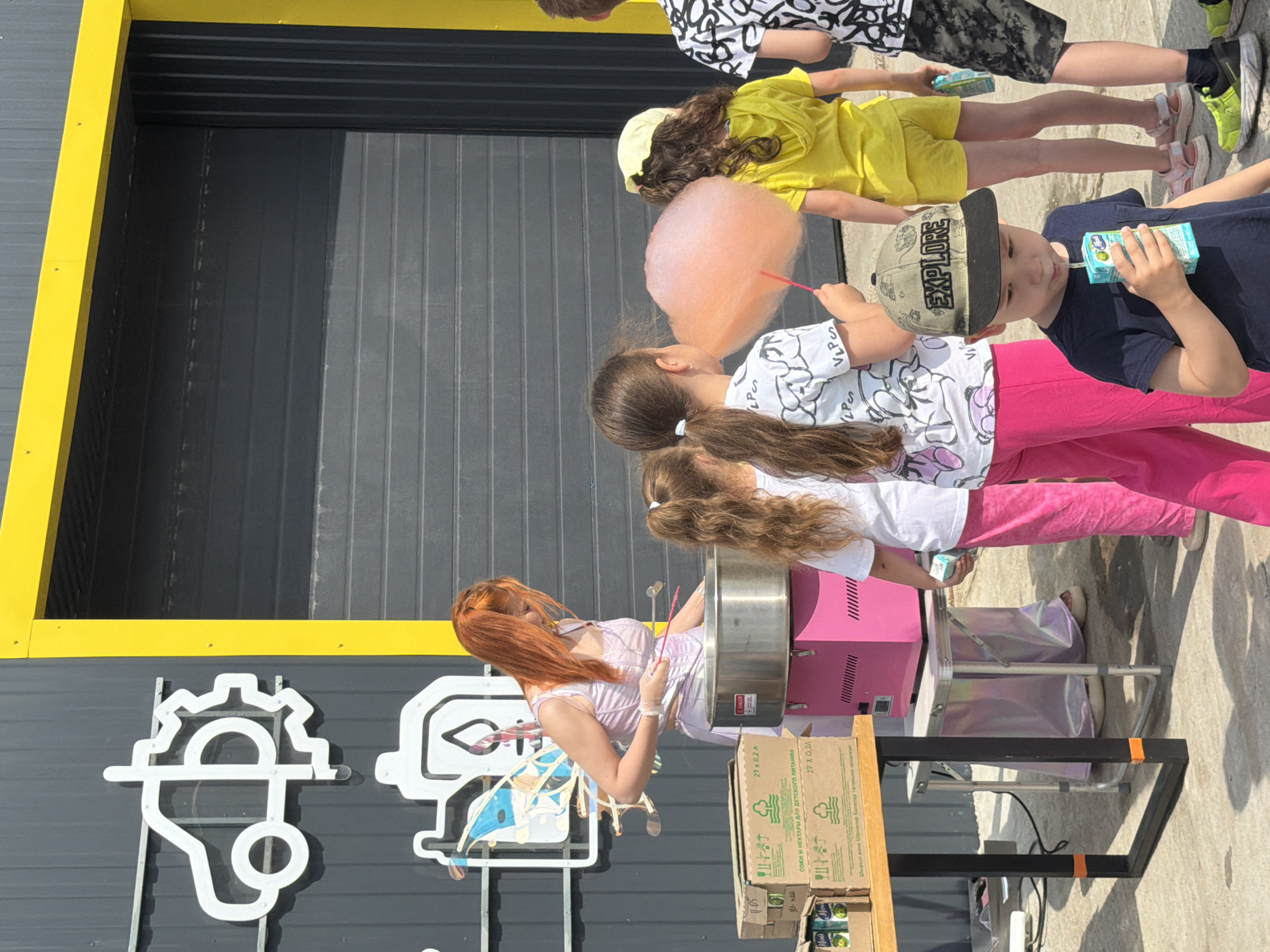 The image depicts an outdoor scene in the city of Chaikovsky, Russia, likely on a sunny day. In the background, a building with a grey corrugated metal facade and a bright yellow framed entrance is visible. There are two stylized white graphic signs mounted on the building's facade, one resembling a car with a gear, and another depicting a container with a flame.
In the foreground, several children and an adult are present. A woman with long red hair, wearing a light purple outfit and what appear to be fairy wings, stands behind a pink cotton candy machine, seemingly in the process of making cotton candy. She holds a stick with a piece of pink cotton candy.
Several children are gathered around, some observing the cotton candy making and others holding drinks. One child, wearing a dark blue t-shirt and a green and black cap with "EXPLORE" written on it, is drinking from a juice box. Another child in a yellow hoodie and shorts is also visible. The overall atmosphere suggests a casual gathering or event, possibly a fair or outdoor party. The ground is paved.