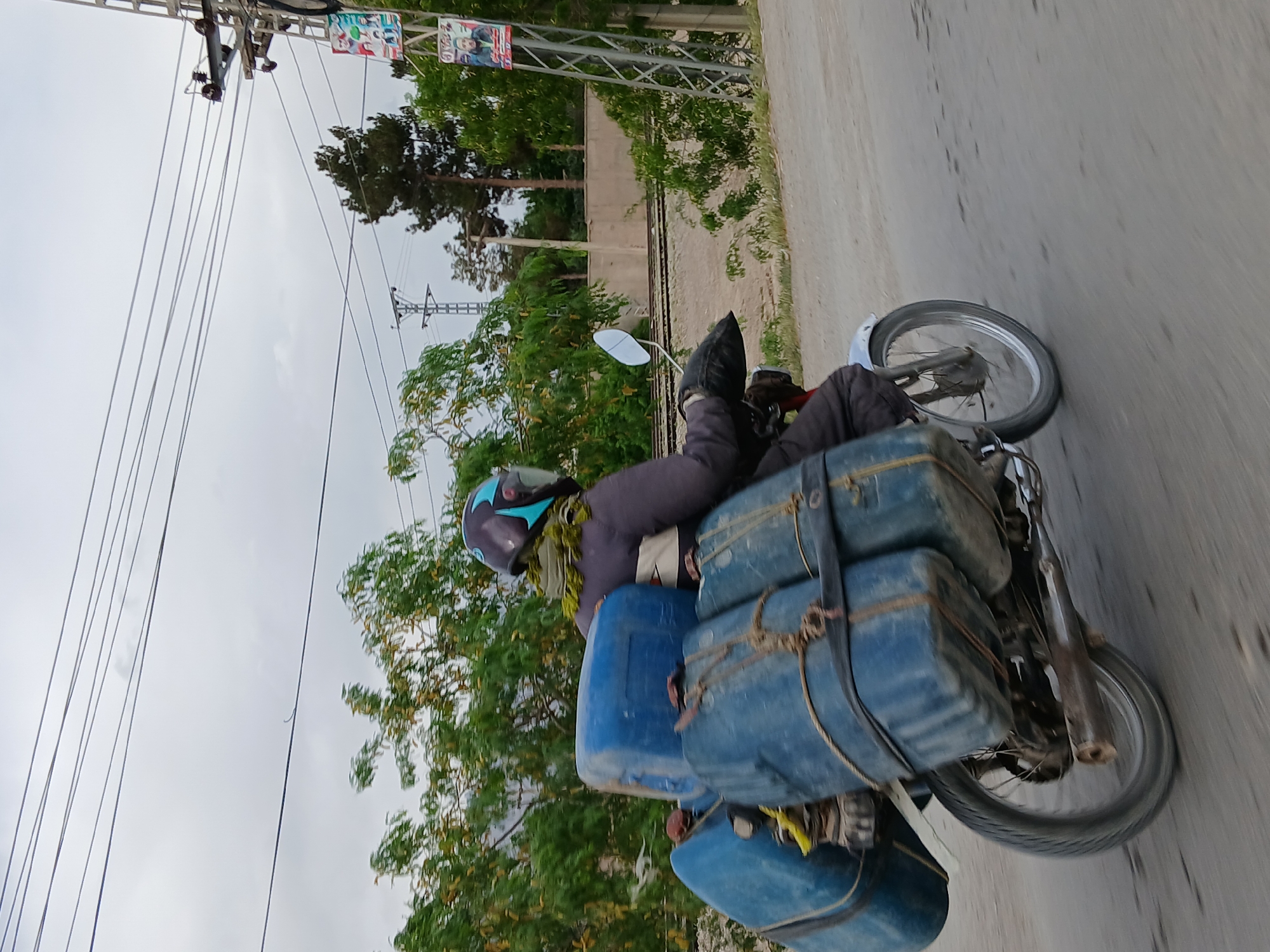 The image shows a motorcycle traveling on a paved road in Quetta City Tehsil, Pakistan. The motorcycle is heavily loaded with blue containers secured with rope and straps. The rider is wearing a helmet and dark clothing. Utility poles with power lines are visible on the left side of the frame. Trees and vegetation line the background, with railway tracks also visible in the distance. The sky is overcast.