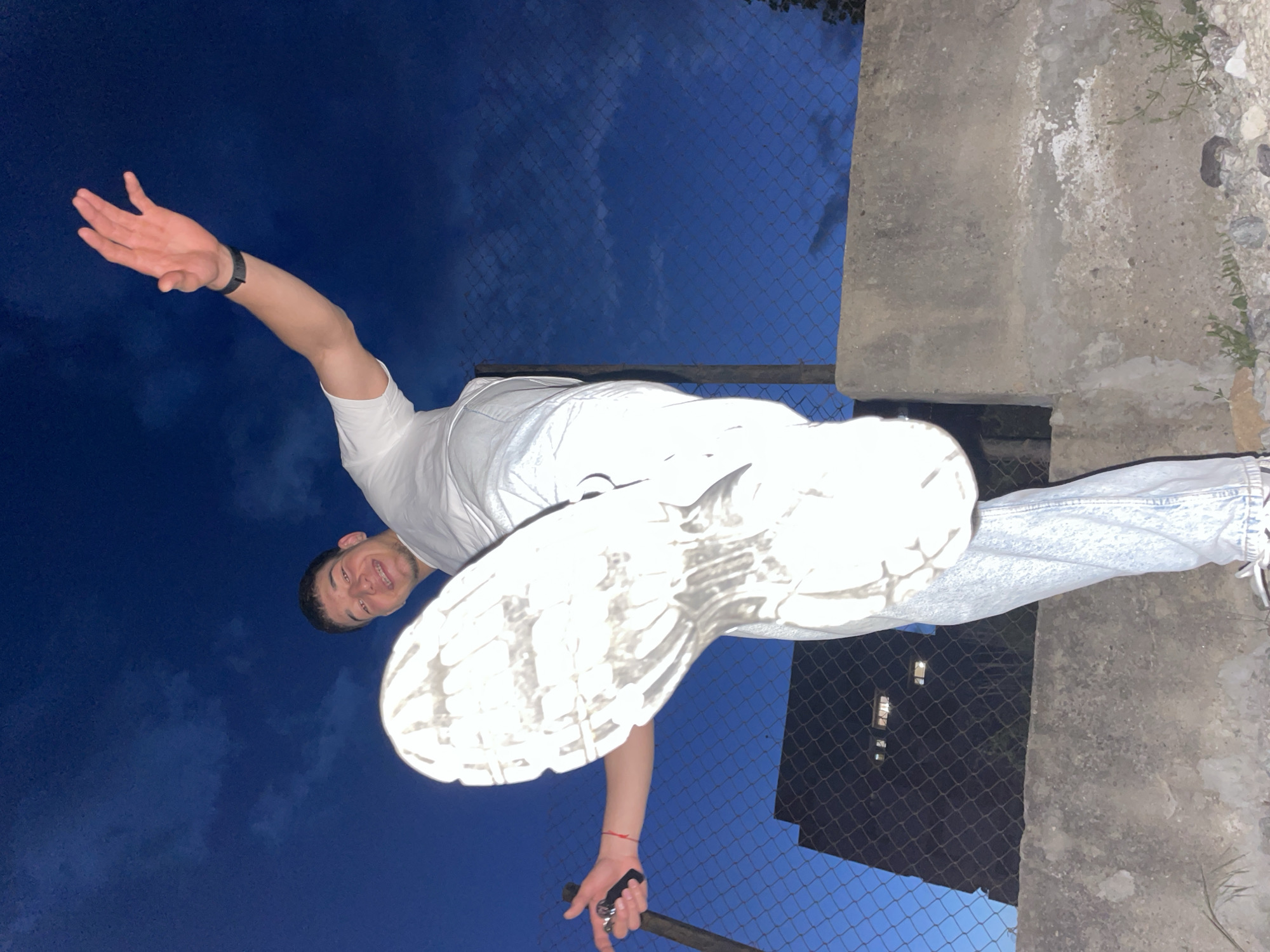 A low-angle shot depicts a man in a white t-shirt and light-wash jeans standing near a concrete structure and a chain-link fence. He extends his left arm upward and outward, while holding a dark object in his right hand. The scene appears to be near an apartment building at night, and is illuminated from the side. The man is smiling. The location is in a city and resembles Zlatograd, Bulgaria.