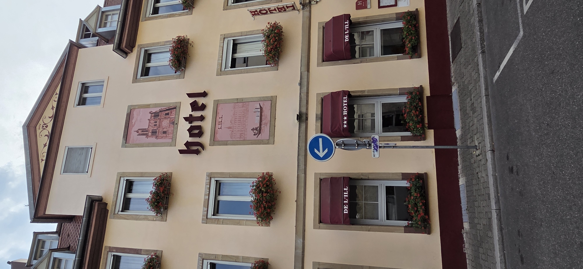 This is a low-angle, full-shot, eye-level, outdoor, daylight, eye-level photograph of the Hotel De L'Ill building located at 6 Rue de la Fontaine in Strasbourg, France. The hotel is a multi-story building with a pale yellow facade and a dark red base. The facade is adorned with several paintings, including one of a cathedral and another depicting a canal scene with a boat. The word "hotel" is prominently displayed in large, dark red letters. Several windows are visible, each framed by brown trim and decorated with flower boxes filled with red flowers. Burgundy awnings extend over some of the windows, with "DE L'ILL" and "*** HOTEL" printed on them. A blue circular sign with a white arrow pointing left, indicating a direction, stands next to the hotel. A street with grey pavement and a white parking line is in the foreground. The sky above is overcast.