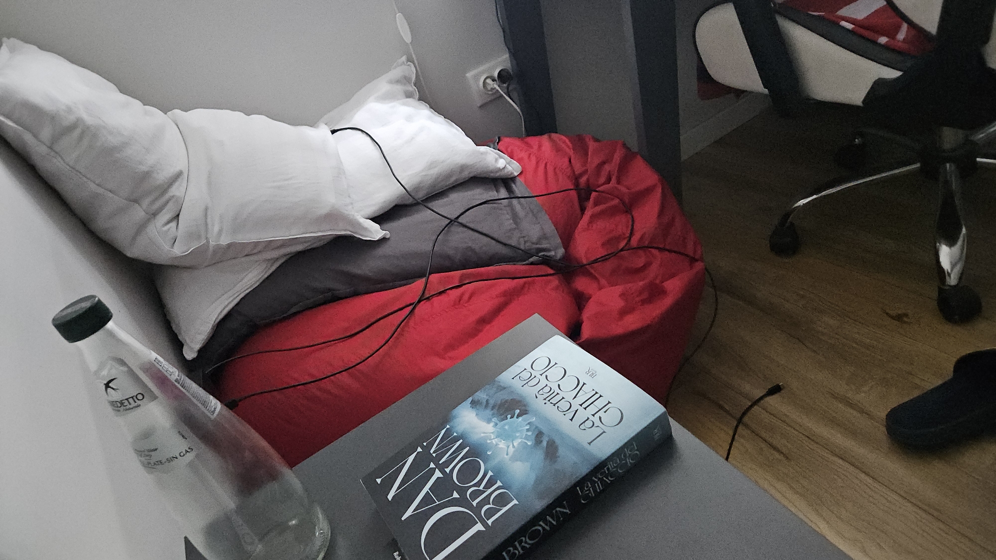 The image presents a cluttered indoor scene. The primary focus is on a stack of pillows, two white and one grey, resting against a wall next to a red beanbag chair. A bottle of water sits on a grey surface, likely a nightstand or small table. On the table is a book titled "La verità del ghiaccio" by Dan Brown. The background includes part of an office chair with a red and white pattern, and a dark slipper on a wooden floor. An electrical outlet and some black cables are also visible. There is no information to suggest this is located in Plovdiv, Bulgaria.