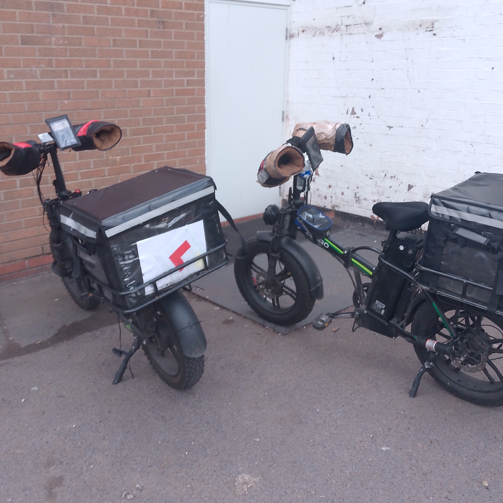 Two electric bicycles are parked on asphalt in front of a brick and white-painted brick building, likely in Coventry, United Kingdom. Both bicycles have black cargo boxes attached to the rear and a red 'L' plate displayed. The handlebar grips are covered with hand mitts, and both bikes are equipped with screens. The bike on the right has a green trim and an additional bag attached to the handlebars. The overall scene suggests the bikes are used for delivery services.