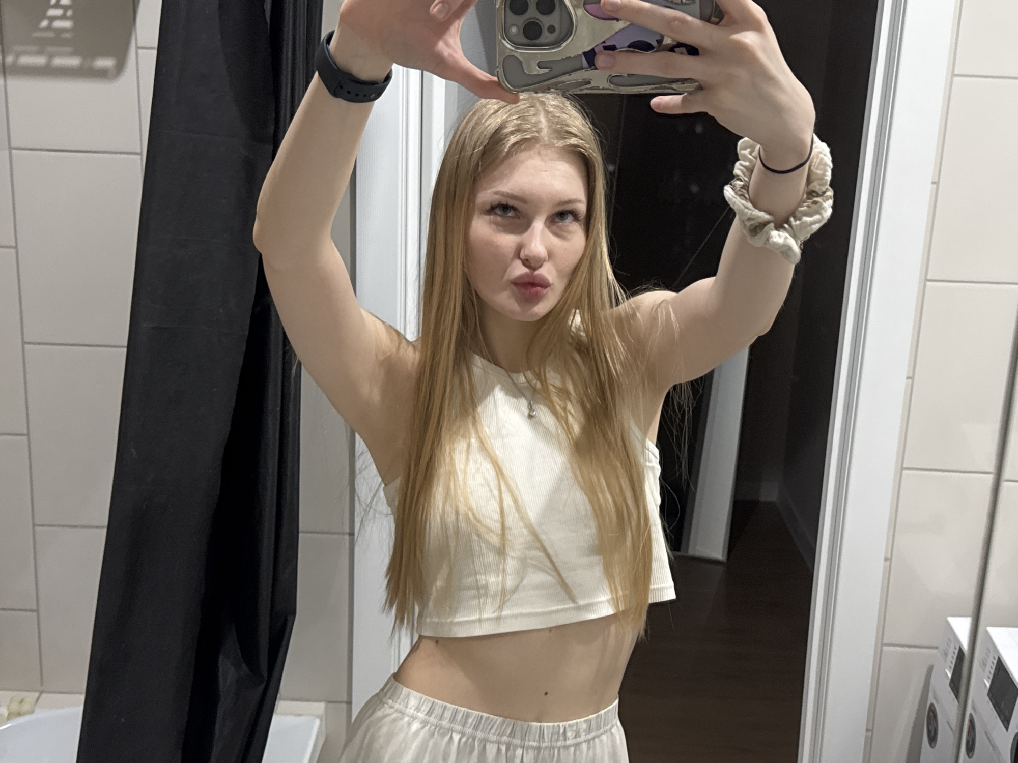 A young woman with long, light blonde hair is taking a selfie in a bathroom. She is wearing a white crop top and light-colored lounge pants. She is holding a phone up in front of her face. She is standing in front of a bathroom mirror, which reflects a doorway leading into another room. A black shower curtain is visible to the left. White tiles cover the wall behind the woman. The city of Perm, Russia, is not discernible in the image.