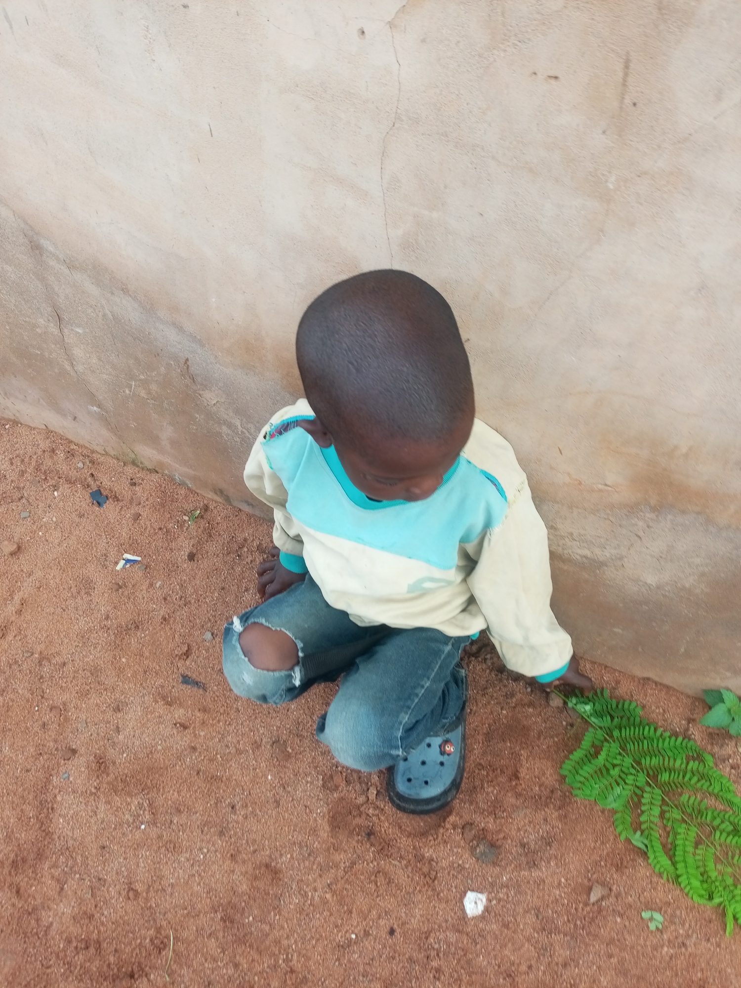 In Tempure, Nigeria, a young child is crouched on reddish-brown dirt near a light beige, textured wall. The child wears a long-sleeved beige shirt with light blue accents on the collar and sleeves, and ripped blue jeans with a tear on the left knee. On their feet are blue Croc-style shoes. To the child's right, a vibrant green fern lies on the ground. Several small pebbles and pieces of debris are scattered on the dirt surface. The scene is captured from a high-angle perspective, looking down on the child.