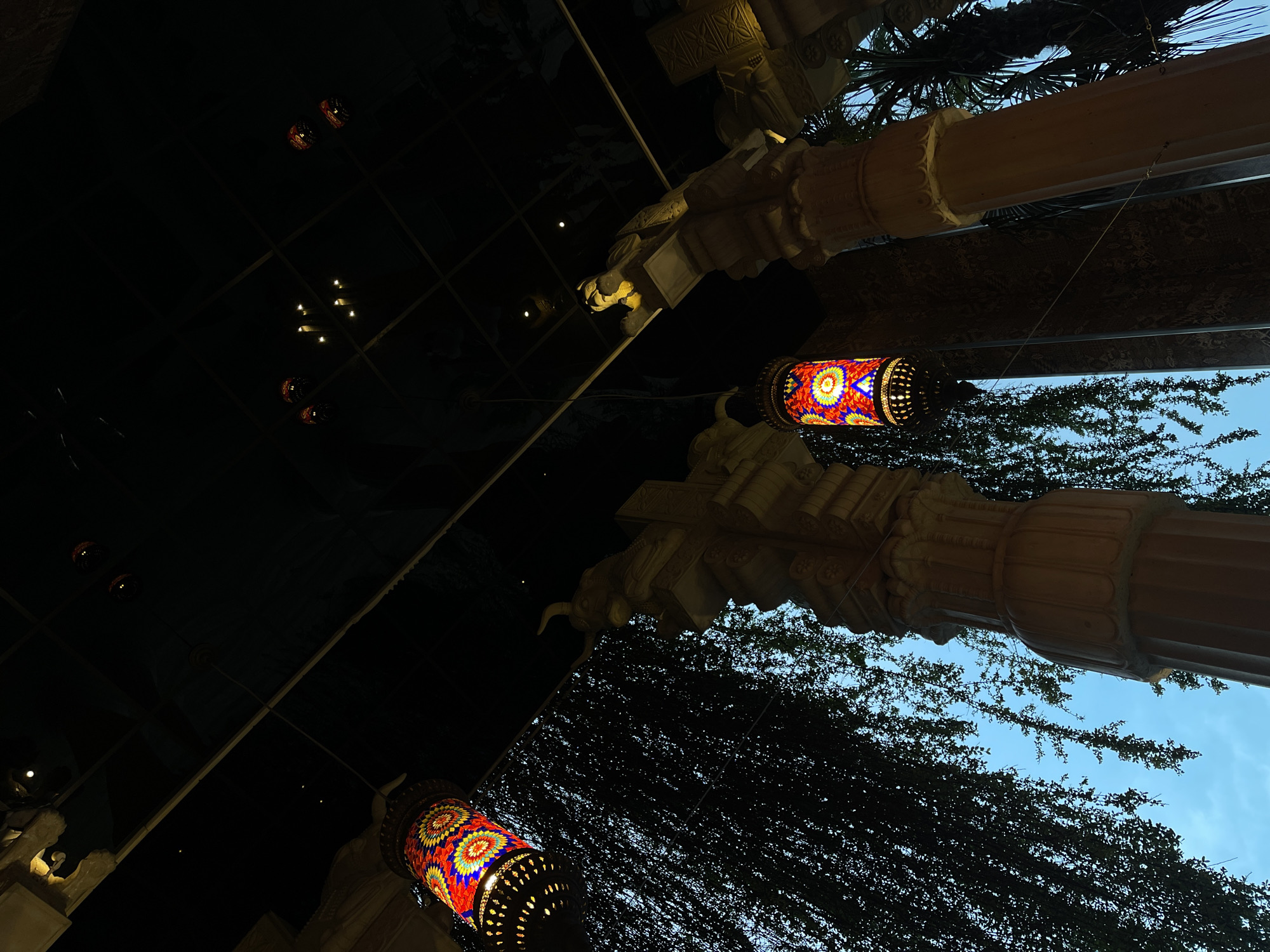 The image captures an upward-looking view of an ornate architectural setting, likely a covered outdoor or semi-enclosed space, at dusk or night. Two vibrant, cylindrical lanterns with intricate mosaic patterns, featuring red, blue, yellow, and green colors, hang prominently. One lantern is in the lower left, and another is in the mid-right, both emitting a warm, inviting glow.
Large, decorative columns or pillars, possibly made of stone or concrete, frame the scene. These columns exhibit detailed carvings and capitals, suggesting a grand and elaborate architectural style. One substantial column dominates the right side of the frame, extending from bottom to top.
A dark, reflective surface, possibly a ceiling or a mirrored wall, occupies the upper left portion of the image. This surface reflects faint grid lines and small, distant points of light, adding depth and a sense of mystique.
Lush green foliage, including dense vines and the fronds of a palm tree, is visible, particularly on the right side and in the background, contrasting with the man-made structures. In the upper right corner, a patch of clear, soft blue sky is visible, confirming the outdoor setting and suggesting it is twilight.
The overall atmosphere is one of exotic elegance and tranquility. The scene is static, showcasing the aesthetic details of the location. No people or visible text are present. Given the context, this ornate setting is located in Varna, Bulgaria.