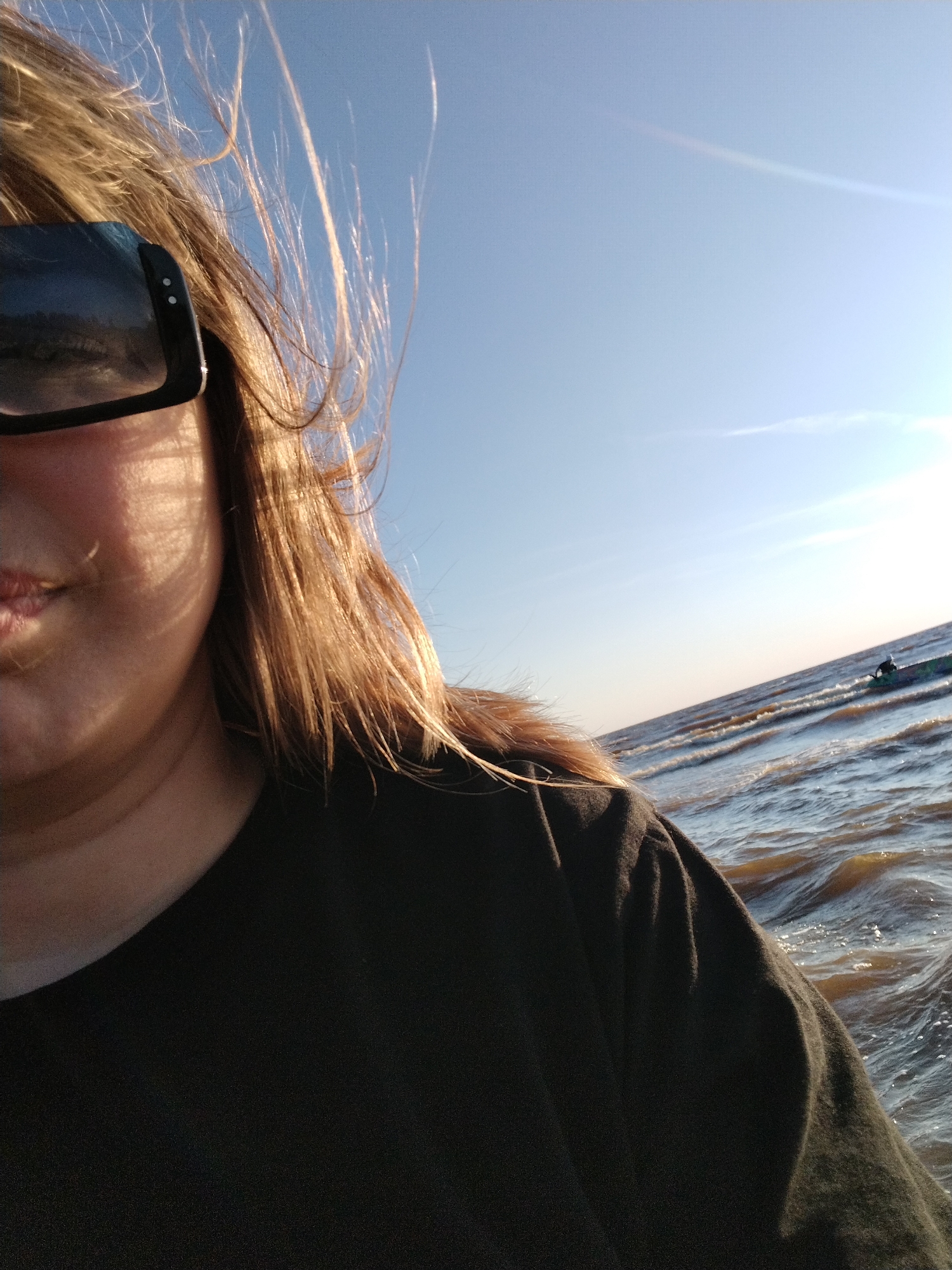 A close-up, outdoor, eye-level selfie shows a person with blonde hair and sunglasses standing by a body of water. The person is wearing a black t-shirt and their face is partially visible on the left side of the frame. Sunlight is reflecting off the water, creating a shimmering effect. The sky is clear and blue, with a hint of sunlight on the right side of the frame. In the distance, a person is visible on a boat in the water. The scene appears to be a sunny day in Ivanovskoe, Russia.