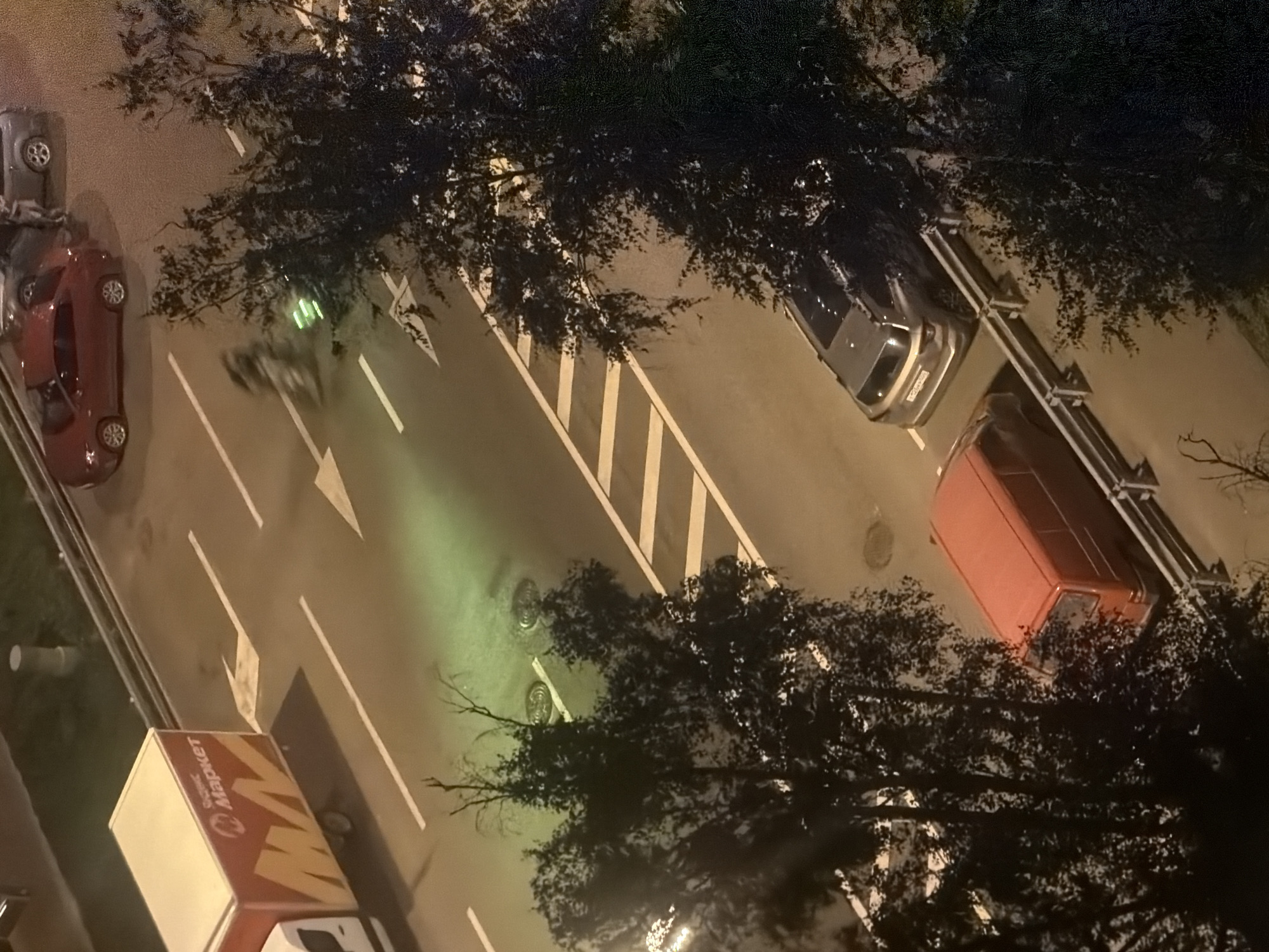 This high-angle aerial view captures a nighttime scene on a paved urban street, likely in Moscow, Russia, given the Cyrillic text on the truck. The street is marked with white lane lines and a pedestrian crossing.
Dominating the left side of the frame is a white truck with a red and yellow cargo box featuring Cyrillic text that reads "СберМаркет" (SberMarket). To the right of the truck, a red sedan is parked. Further down the street, a silver SUV is parked with its rear facing the camera. A bright red van is also visible parked on the right side of the street.
In the middle of the street, there is a blurry, green illuminated object that appears to be a drone or a similar aerial device. Several trees with dark foliage are scattered throughout the scene, obscuring parts of the view, particularly on the right side. The lighting suggests artificial streetlights and vehicle headlights illuminating the area. Two manhole covers are visible on the street surface.