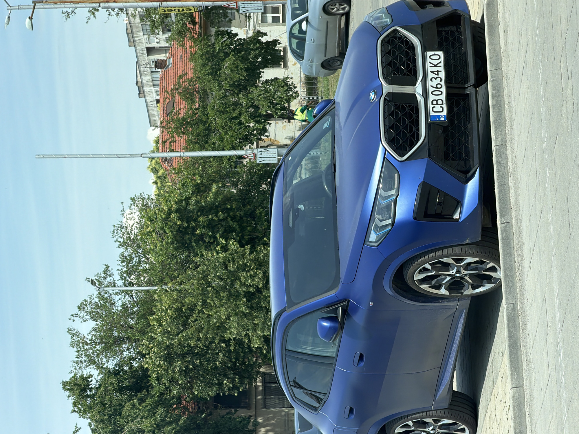 A vibrant blue BMW X1 (likely U11 generation) is prominently parked on the right side of a sunlit urban street in Plovdiv, Bulgaria, facing left. The car's front fascia, featuring its distinctive large black kidney grille, modern LED headlights, and multi-spoke alloy wheels, is clearly visible. A Bulgarian license plate, "CB 0634 KO," is affixed to the front, indicating registration in Sofia.
Behind the blue BMW, a silver hatchback, possibly a SEAT Ibiza, is also parked. Further back, partially obscured by trees, a person wearing a bright yellow or green high-visibility vest can be seen. The street is lined with lush green trees, suggesting a pleasant daytime setting. In the background, various buildings with light-colored facades and reddish-brown tiled roofs are visible under a clear, bright blue sky. Utility poles or streetlights extend upwards throughout the scene. A yellow sign with partially visible, reversed text, possibly "erbet" or similar, hints at a commercial establishment. The overall impression is a peaceful street scene on a clear, sunny day.