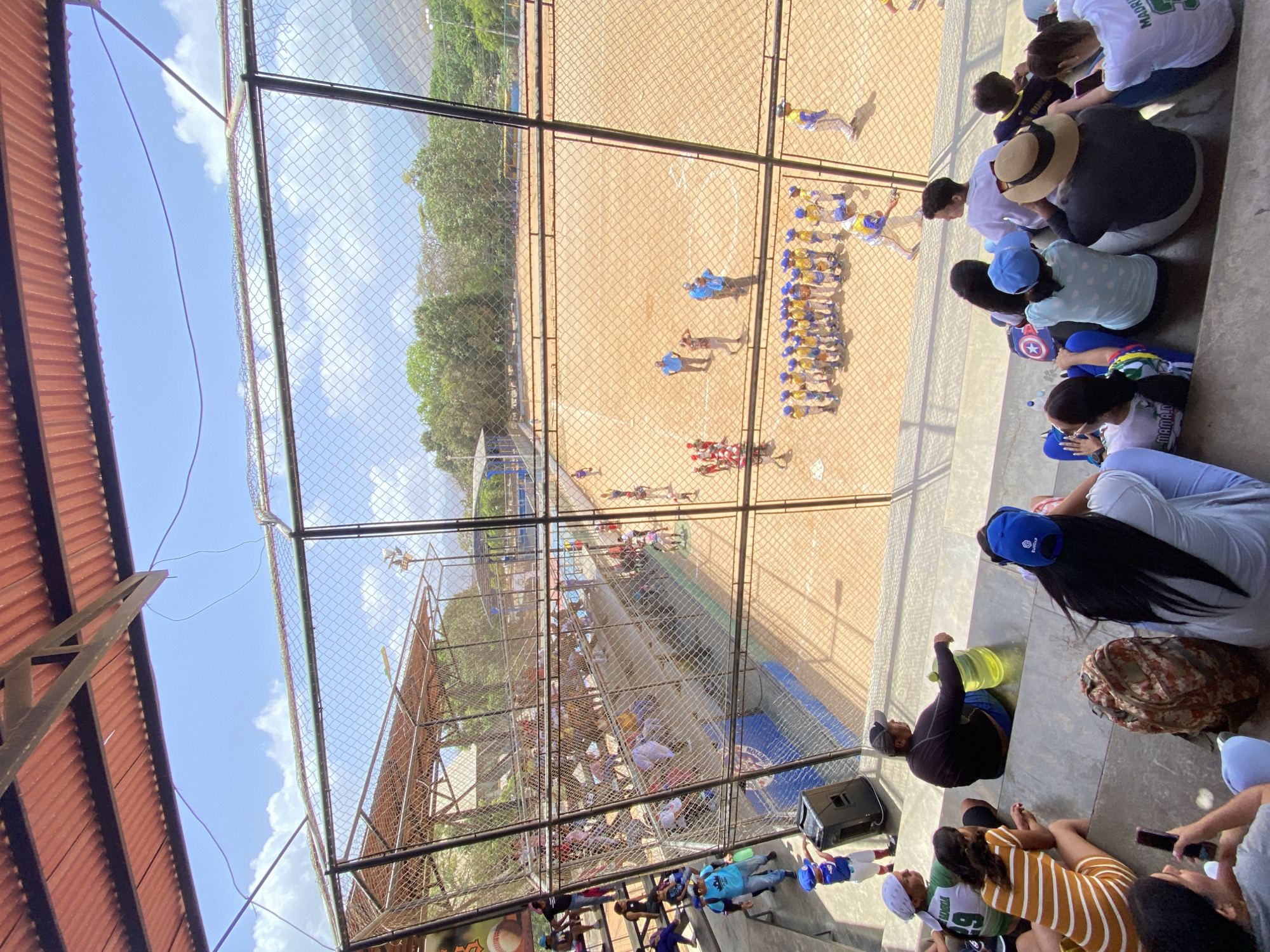 The image shows a baseball field in La Asuncion, Venezuela, viewed from an elevated seating area. A chain-link fence separates the seating from the field. On the field, two groups of baseball players, one in blue and yellow uniforms and the other in red uniforms, are interacting. The seating area is filled with spectators. The sky is visible above the structure, with some clouds present.