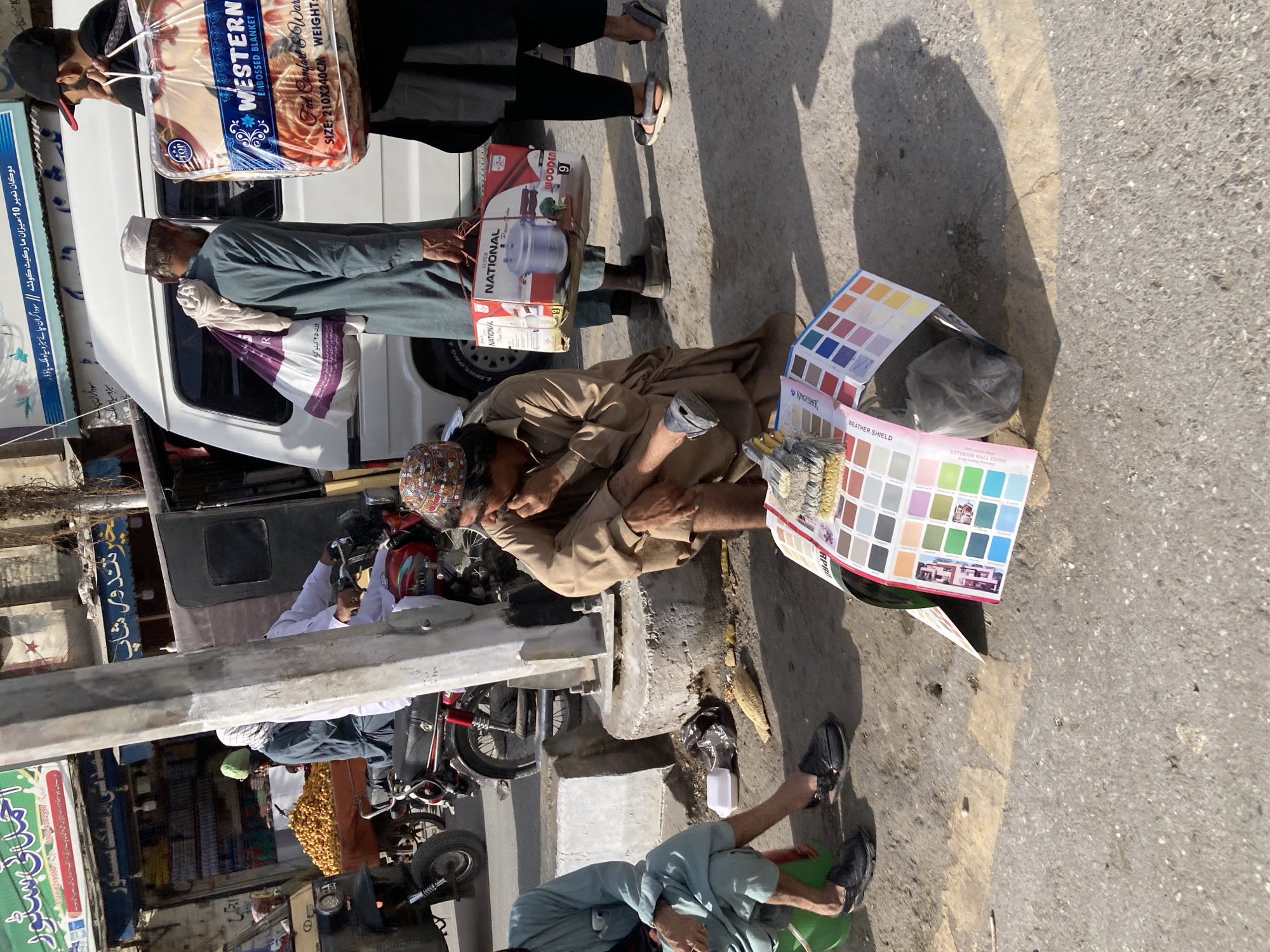 This image captures a bustling street scene in Quetta City Tehsil, Pakistan, under bright daylight, suggested by the strong shadows. The setting appears to be a market or commercial area with shops lining the street.
Several individuals are visible. In the foreground, a man in a dark cap and shirt carries a large, clear plastic package containing a "WESTERN EMBOSSED BLANKET" labeled "Fed Comfort & Warmth," sized "210X240CM." Behind him, another man in a white cap and green/grey shalwar kameez stands by a white van, holding a white plastic bag. To their right, a man in a light brown shalwar kameez and a patterned cap is seated on the ground, appearing to be resting or sleeping. Nearby, paint color swatch cards from "KINGFISHER" for "WEATHER SHIELD" and "EXTERNAL WALL FINISH" are laid out, along with paintbrushes, suggesting a vendor or painter.
Further in the foreground, another man in a light blue shalwar kameez and black sandals sits on the ground next to a green bucket. In the mid-ground, a white van is parked, and a man is seen carrying a box for a "NATIONAL WOODEN 9" item. The background reveals partial views of buildings with various shop signs in Urdu, including "Shop No. 10, Mizan Market Quetta // Traders of green and black tea, milk powder," "Traders of tea," and "Khair Tandoor Shop," confirming the location and commercial nature of the area. Motorcycles and a concrete barrier are also part of the street furniture. The scene depicts everyday life and commerce in a vibrant urban environment.