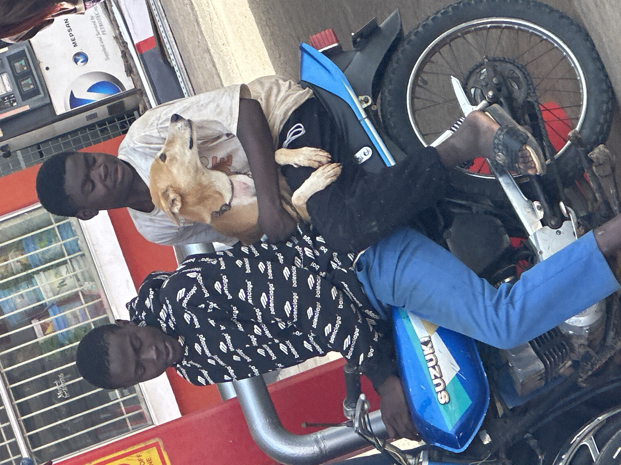 Two young Black males are seated on a blue Suzuki motorcycle in front of a gas station in Gura Top, Nigeria. The male in front, wearing a black hoodie with a repeating white "Balenciaga" logo pattern and blue pants, is holding the handlebars. The male behind him, wearing a light-colored t-shirt and dark pants, is holding a light brown dog. The dog is facing to the right. The gas station appears to be MEPSAN branded. The environment is outdoors with a paved surface.