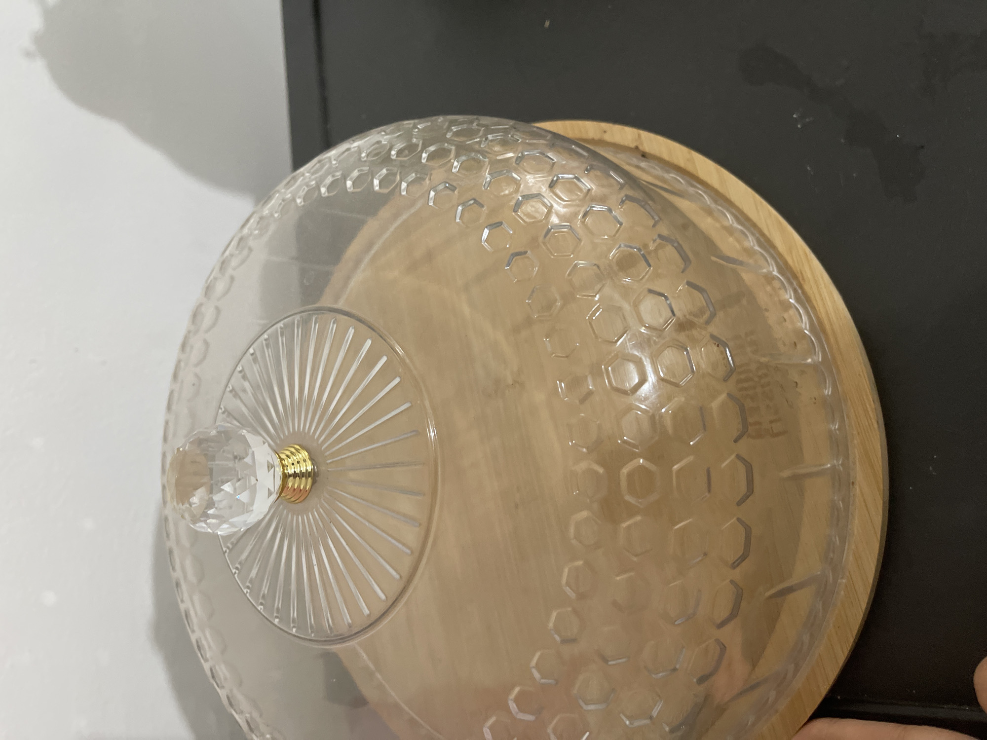 A clear plastic dome with a faceted glass knob sits atop a light-colored wooden circular base. The dome has a decorative embossed honeycomb pattern and radial lines emanating from the knob. The base appears to be a serving platter. The objects are positioned on a dark, flat surface, likely a countertop, with a light-colored wall in the background. A shadow is cast on the wall. The scene appears to be indoors, possibly in a kitchen or dining area. The location is not specified within Bishkek City, Kyrgyzstan.