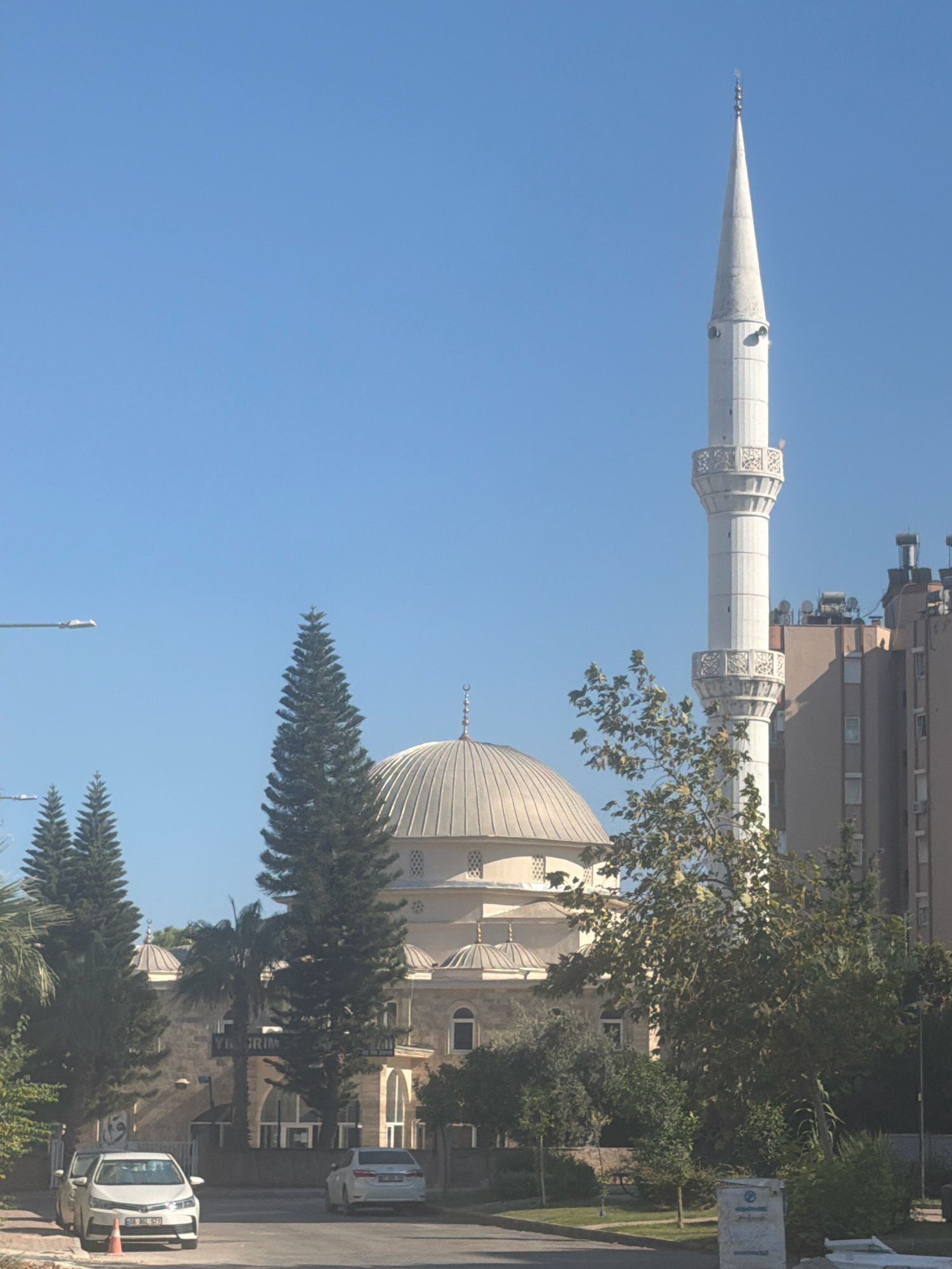 The image depicts the exterior of a mosque and its minaret under a clear blue sky in Antalya, Turkey. The mosque features a prominent dome and several smaller domes, constructed with a light-colored stone. The minaret is tall and slender, made of white material, with decorative balconies. To the right of the mosque, a modern apartment building with several stories is visible. In the foreground, a street is present with two white cars parked on the right side. Several trees, including tall evergreen trees and leafy deciduous trees, are scattered throughout the scene, some obscuring parts of the mosque and buildings. A street lamp is also visible on the left side of the image. The overall environment suggests an urban setting in Antalya, likely in a residential or mixed-use area.