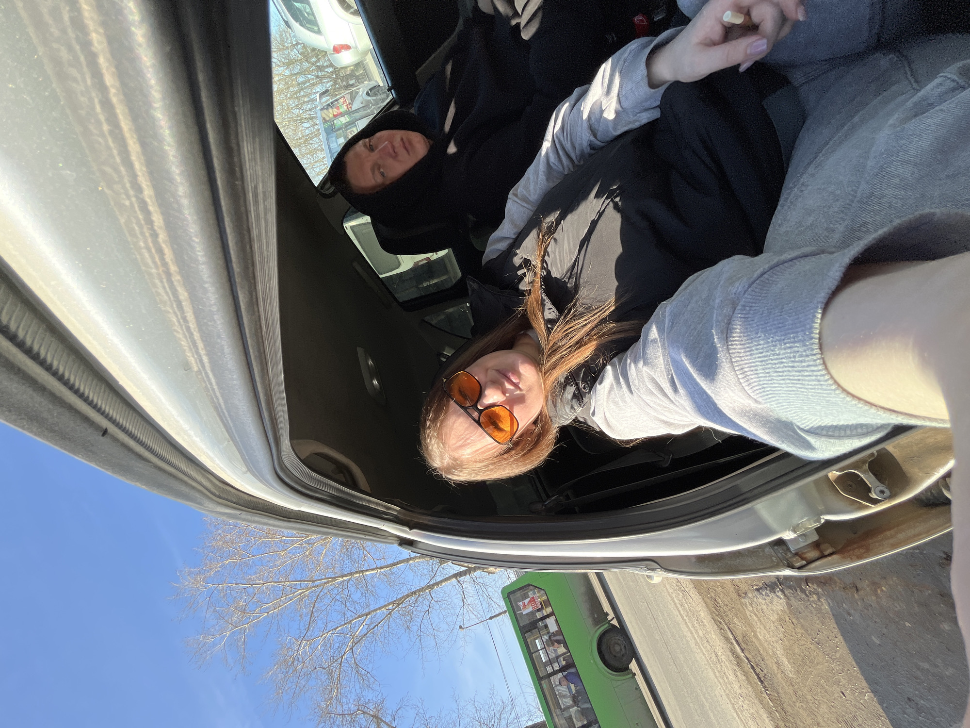 A daylight selfie is captured from the front passenger seat of a light-colored vehicle, parked on a dirt shoulder adjacent to an asphalt road in Chaikovsky, Russia. The front passenger door of the vehicle is open. In the foreground, a woman with long reddish-brown hair wears orange sunglasses, a grey long-sleeved shirt, and a black vest. She holds what appears to be an unlit cigarette in her left hand, while her right arm is extended to operate the camera. In the back seat on the passenger side, a man with a neutral expression is seated, wearing a black hooded sweatshirt with the hood up. Through the open door and windows, a green public transit bus with visible passengers is on the road, alongside bare deciduous trees and a clear blue sky. A light-colored SUV is partially visible parked further down the road.