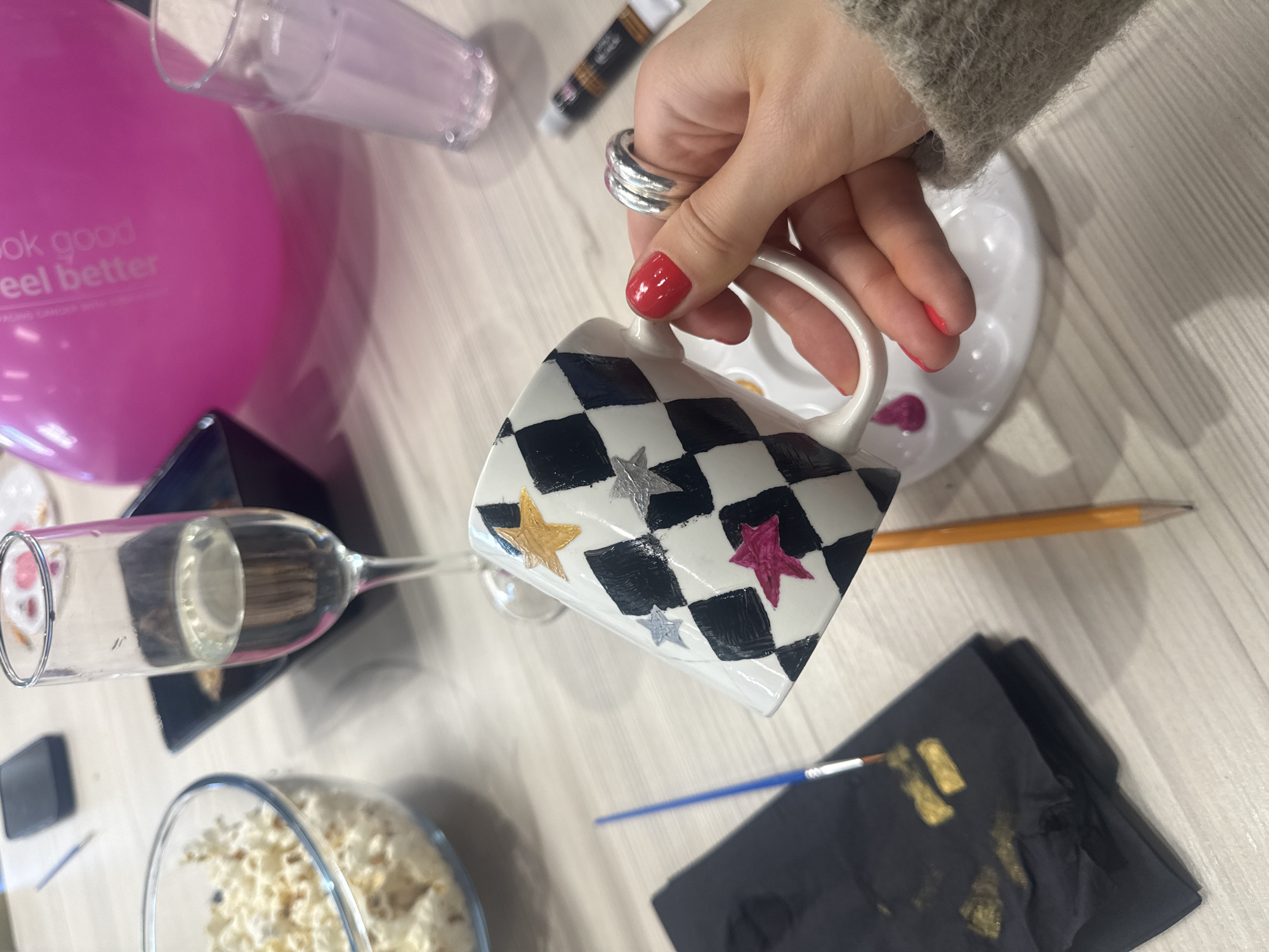 The scene is a well-lit, close-up shot of a table or surface with various objects suggesting a creative activity, possibly painting or crafting. A hand, adorned with two silver rings on the index finger and red nail polish, holds a white ceramic mug decorated with a black and white checkerboard pattern and star motifs of different colors.
Various items are arranged on the table: a pink balloon with text, a tube of paint, a white paint palette, a pencil, a black cloth with what appears to be gold paint smudges, a blue paintbrush, a clear glass bowl filled with popcorn, and an overturned wine glass on a black stand.
The setting is likely indoors. There is no discernible location in the city of London, United Kingdom.