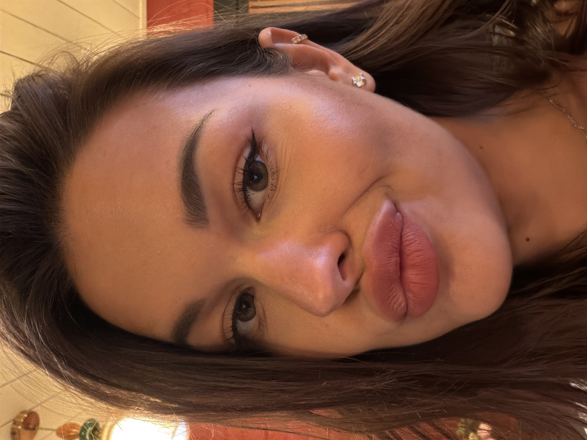 This is a close-up, selfie-style portrait of a young woman with dark brown hair and brown eyes. She is wearing makeup, including eyeliner and lipstick. She has a stud earring in her left ear and a cuff earring on her helix. Her lips are pursed. The background is blurred, but appears to be an indoor setting with warm lighting and some decorative elements visible. There are no discernible identifying features that would suggest a specific location within Saint Petersburg, Russia.