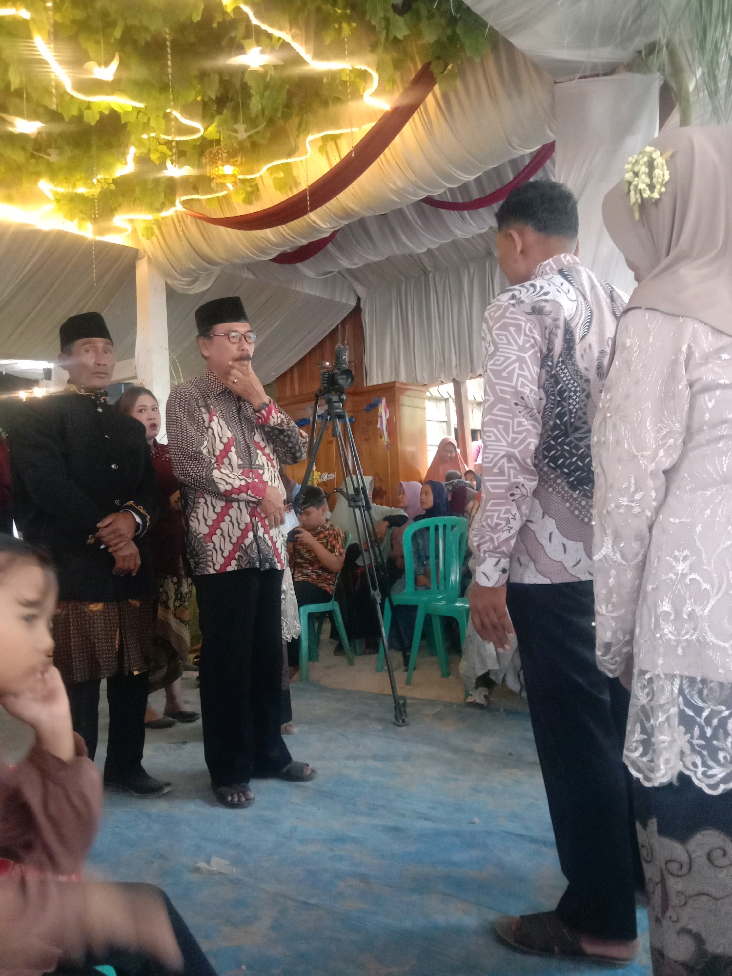 This image captures an indoor gathering, likely a wedding ceremony or reception, in Rejosari, Indonesia. The scene is decorated with white draped fabric and illuminated by warm, yellow string lights intertwined with artificial green foliage overhead.
In the foreground, a child in a brown outfit is partially visible, looking to the left with their hand near their face.
The main subjects are several individuals dressed in traditional Indonesian attire. On the left, a man in a black peci hat and a black traditional jacket with gold trim stands with his hands clasped. Next to him, a man wearing glasses and a patterned shirt with a peci hat is observing something with his hand to his chin. To the right, a woman in a beige hijab with a floral embellishment and a heavily embroidered white dress is visible, standing partially turned away from the camera. Another man in a patterned long-sleeved shirt stands beside her, also turned away.
In the background, other guests are present. A video camera on a tripod is positioned to the right of the central figures. Several green chairs are visible, some occupied by attendees. Wooden furniture and architectural elements of the building are also discernible. The floor is a light blue, possibly concrete.