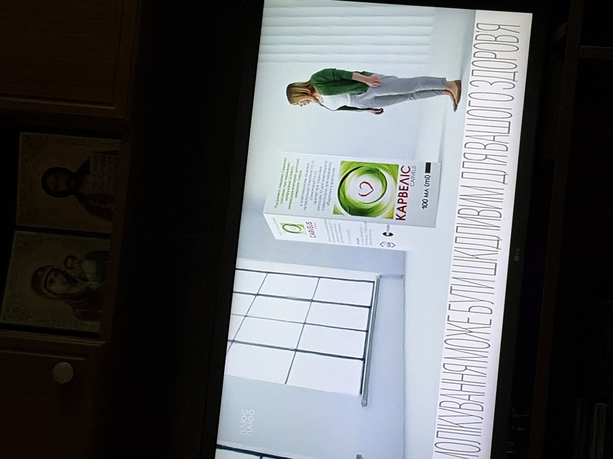 The image shows a television screen displaying an advertisement. In the foreground, text in Cyrillic script reads "МОЛІКУВАННЯ МОЖЕ БУТИ ШКІДЛИВИМ ДЛЯ ВАШОГО ЗДОРОВ'Я". To the left of the text, on the screen, a white box with the logo and name "КАРВЕЛІС" (CARVELIS) is visible. A woman in a green cardigan and white top is standing to the right of the box, looking towards it. The background of the advertisement shows a brightly lit room with a large window. Above the television screen, two religious icons are displayed.