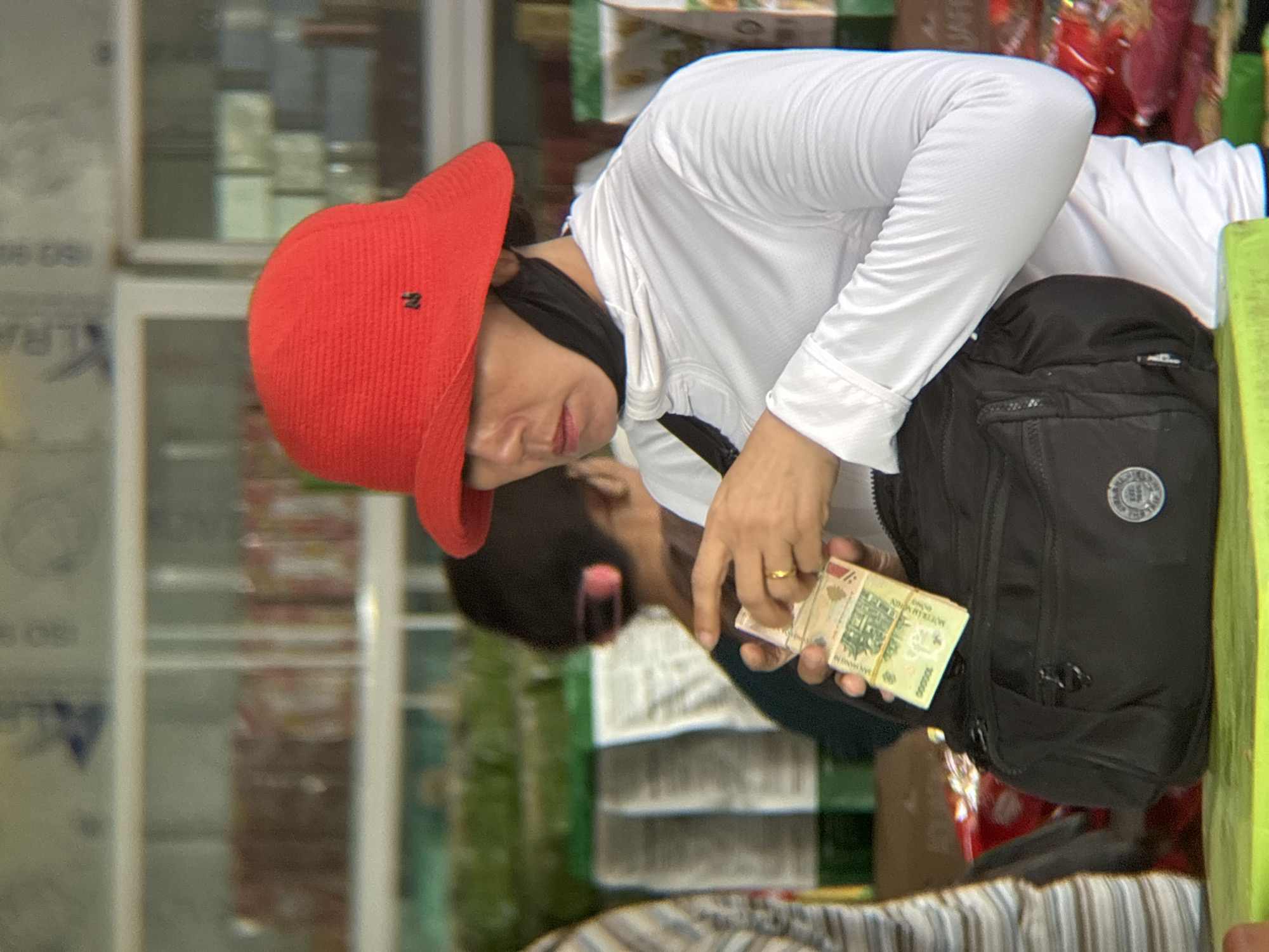 In Lao Bảo Commune, Vietnam, a person wearing a red bucket hat and a white hoodie is holding a wad of currency, possibly Vietnamese Dong, and placing it into a black fanny pack. The person is also wearing a black face mask around their neck. The background appears to be a market or store setting with shelves stocked with goods.