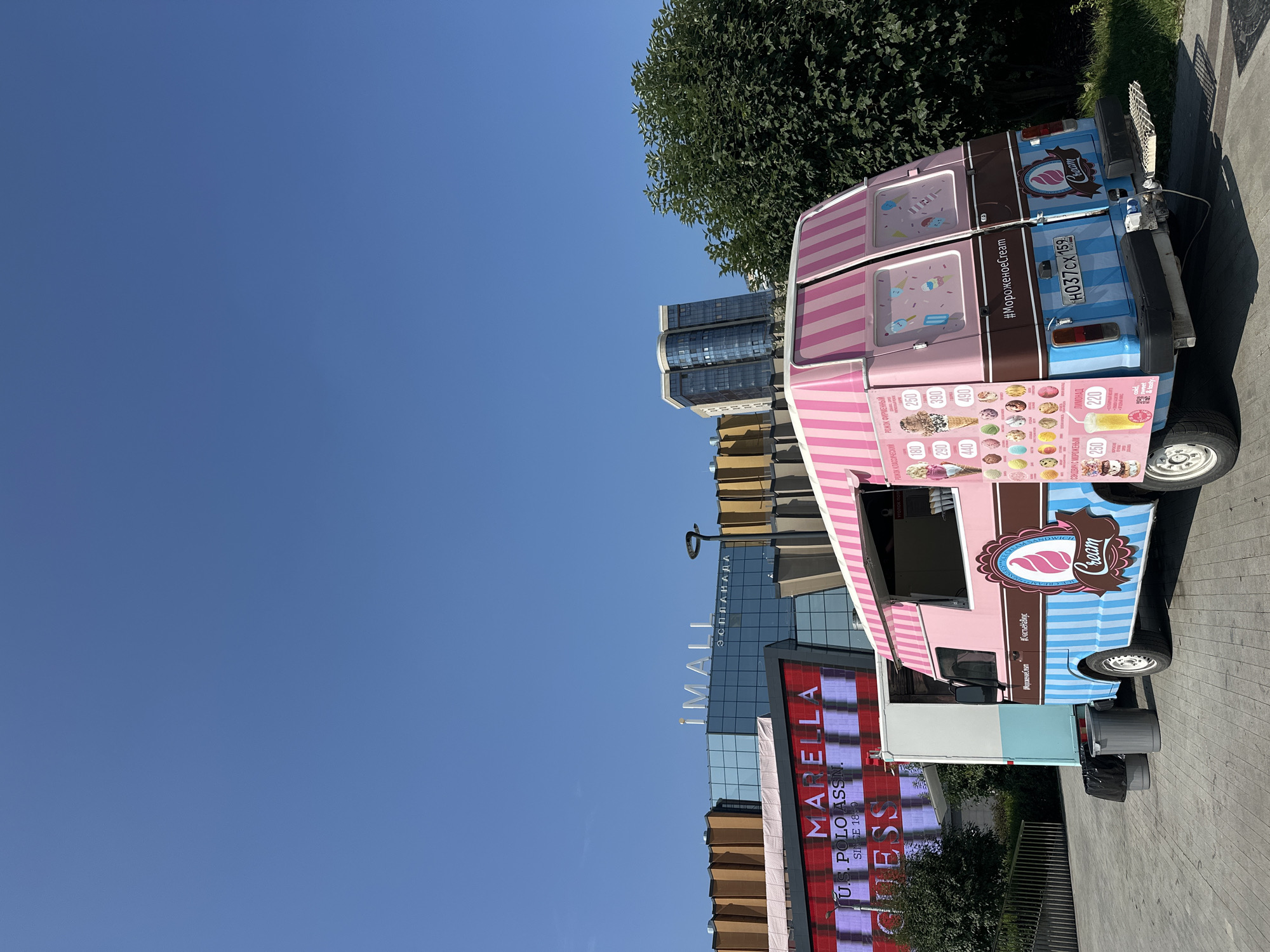 An ice cream truck, painted pink and blue with striped accents and adorned with ice cream graphics, is parked in an urban outdoor setting under a clear blue sky. The truck is the primary subject, positioned in the foreground and facing towards the right of the frame. The rear of the truck displays a price list for various ice cream flavors and options, with text in Russian. A license plate reading "H037CX 159 RUS" is visible.
In the background, a modern shopping mall with glass facades and distinctive architectural features, including a large digital billboard displaying "MARELLA," "U.S. POLO ASSN.," and "GUESS," is visible. The mall appears to be the iMALL shopping center, located in the city of Perm, Russia. Further in the background, a tall, contemporary building with a glass facade stands amongst lush green trees. The ground is paved, suggesting a public area or plaza. Two gray trash cans are placed near the left side of the ice cream truck. No people are present in the image.