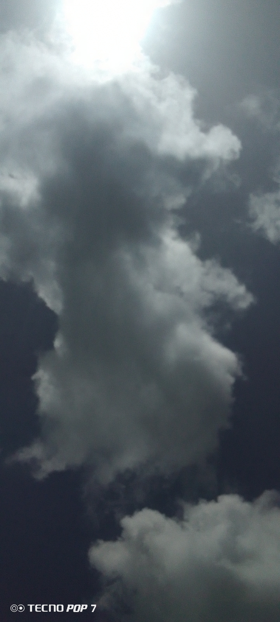 A low-angle, medium shot shows a sky filled with a mix of light and dark gray clouds. Sunlight is visible at the top of the frame, behind some clouds. The overall impression is of an overcast day. At the bottom left of the image are the words "Tecno Pop 7". It's impossible to tell the location in Pedro González, Venezuela from the given image.