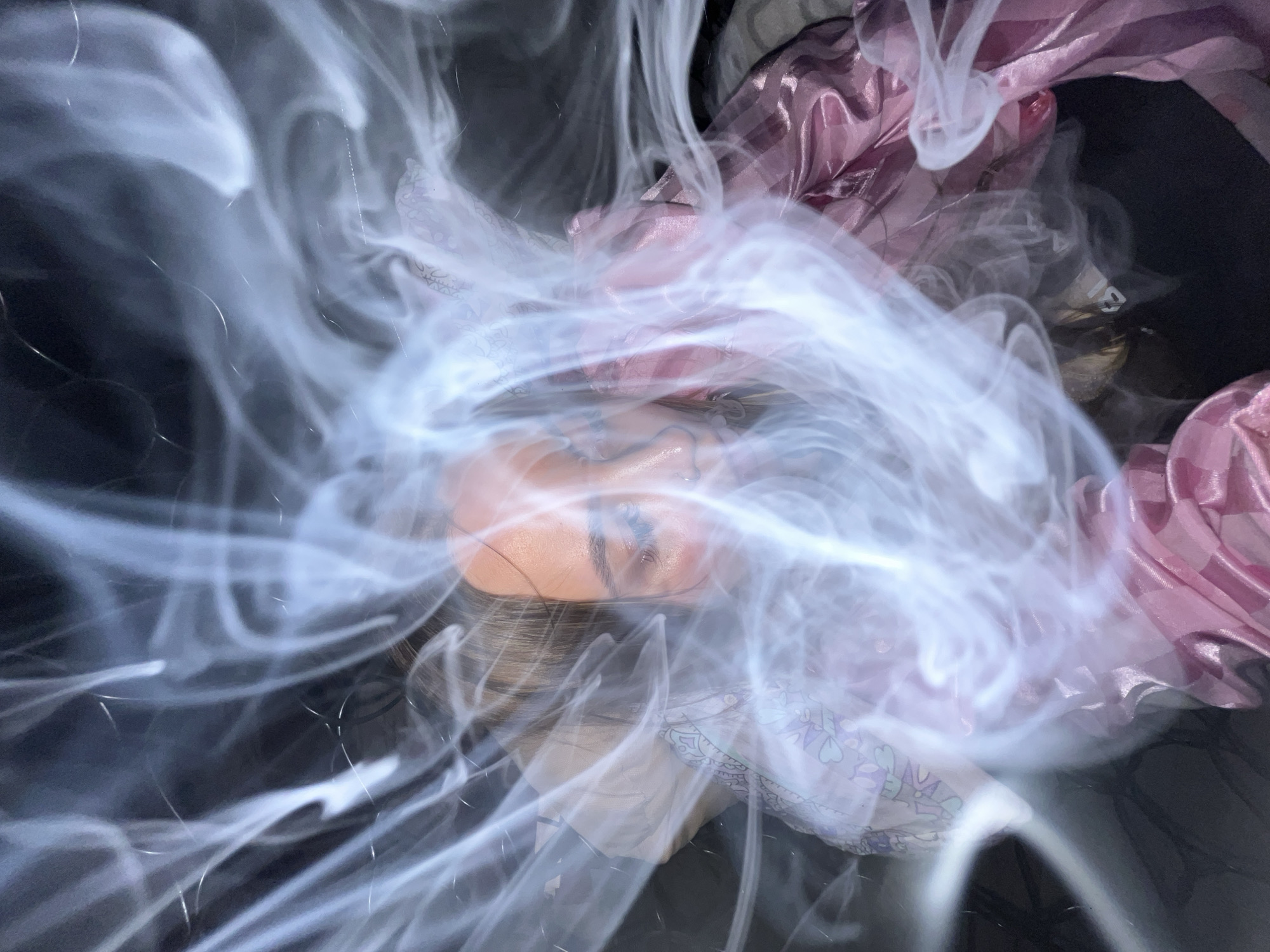 The image presents a close-up, obscured view of a person's face, specifically the forehead and closed eyes. Swirls of light, translucent material resembling smoke or fabric partially cover the face, creating a hazy effect. The person has dark eyebrows and appears to be lying down. In the background, there are pieces of pink and white patterned fabric, suggesting a soft or draped setting. The surrounding environment is dark and blurry, making it difficult to discern further details. There is no indication of the location being Novopokrovskaya, Russia.