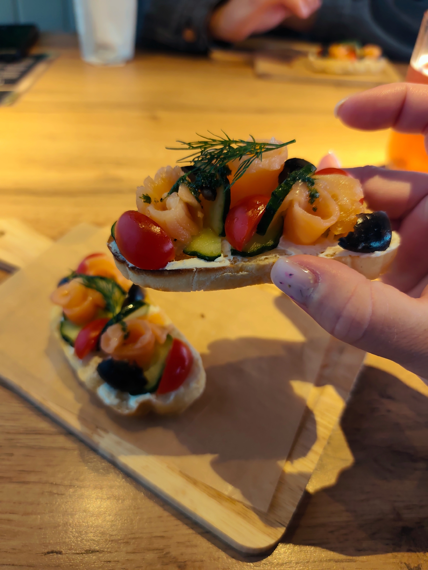 A person is holding a canapé topped with smoked salmon, sliced cucumber, cherry tomatoes, olives, dill, and a creamy spread. Another canapé of similar composition is on a wooden serving board on a wooden table. The location is potentially a restaurant or home setting. No further details confirm the city of Chaikovsky, Russia.