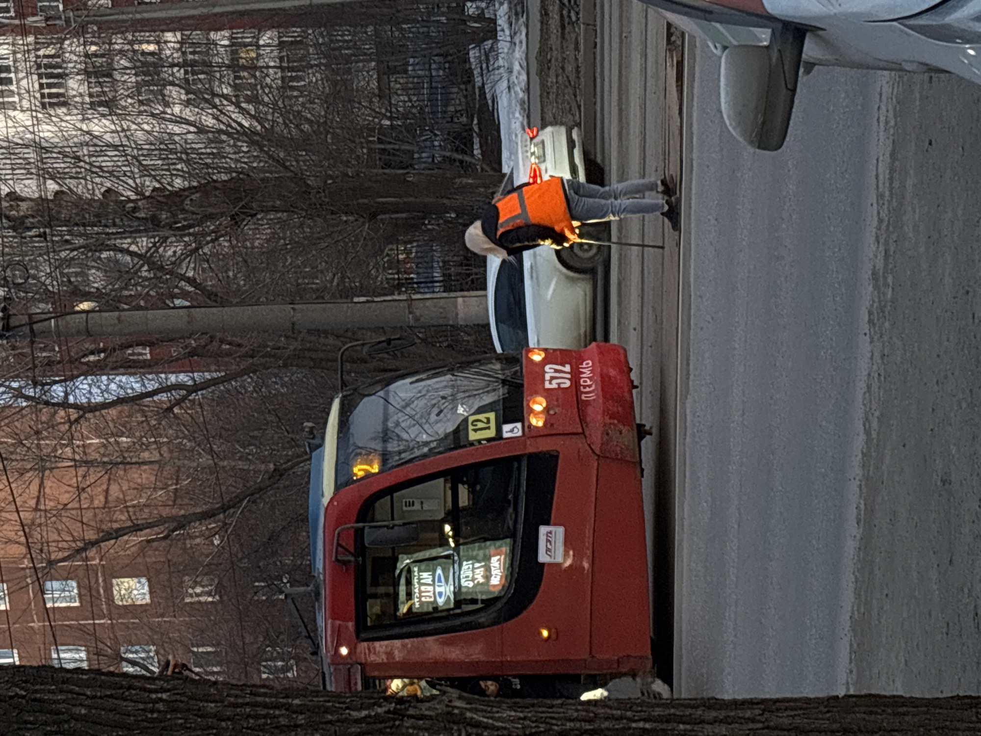 The image shows a red trolley bus with the number "572" and the word "Пермь" (Perm) on its side, which indicates it is located in the city of Perm, Russia. A worker in an orange safety vest is using a long pole to adjust or repair the overhead electrical lines for the trolley bus. A white car is parked nearby. The surrounding environment includes bare trees, a building in the background, and an urban street scene.