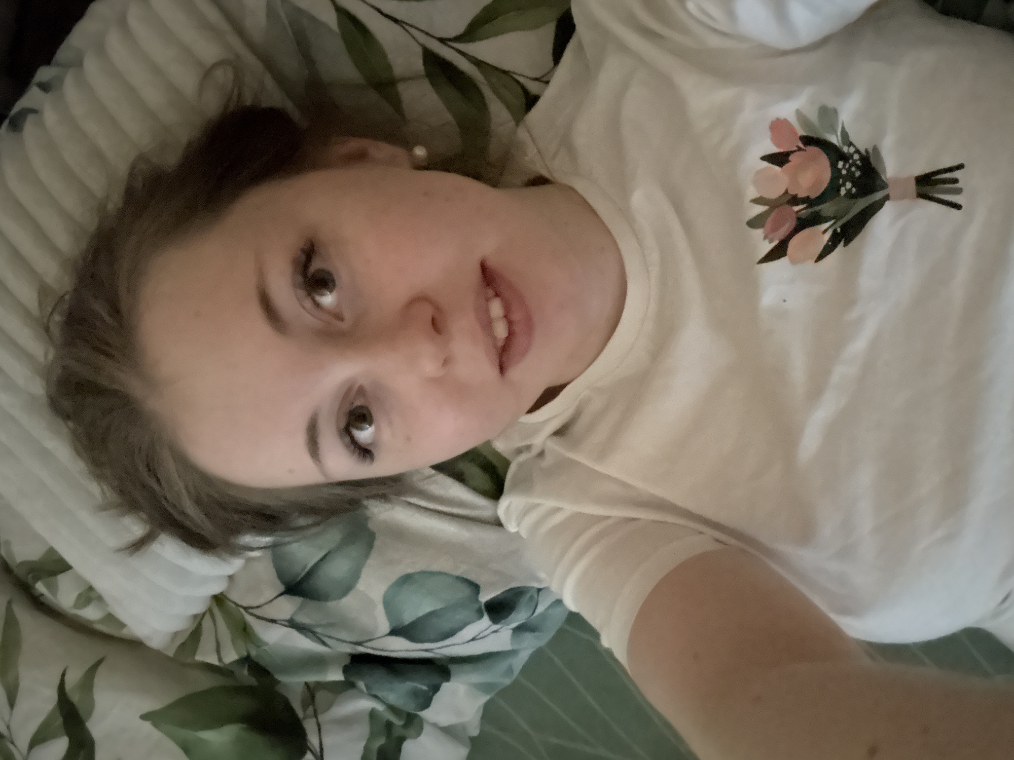 A woman is lying down. She is wearing a white t-shirt with a floral design on the right breast. She is lying on a bed with patterned sheets and a textured pillow. Her head is on the left side of the frame. Her hair is light brown. She is looking at the camera and smiling slightly. The location is Unknown, Unknown.