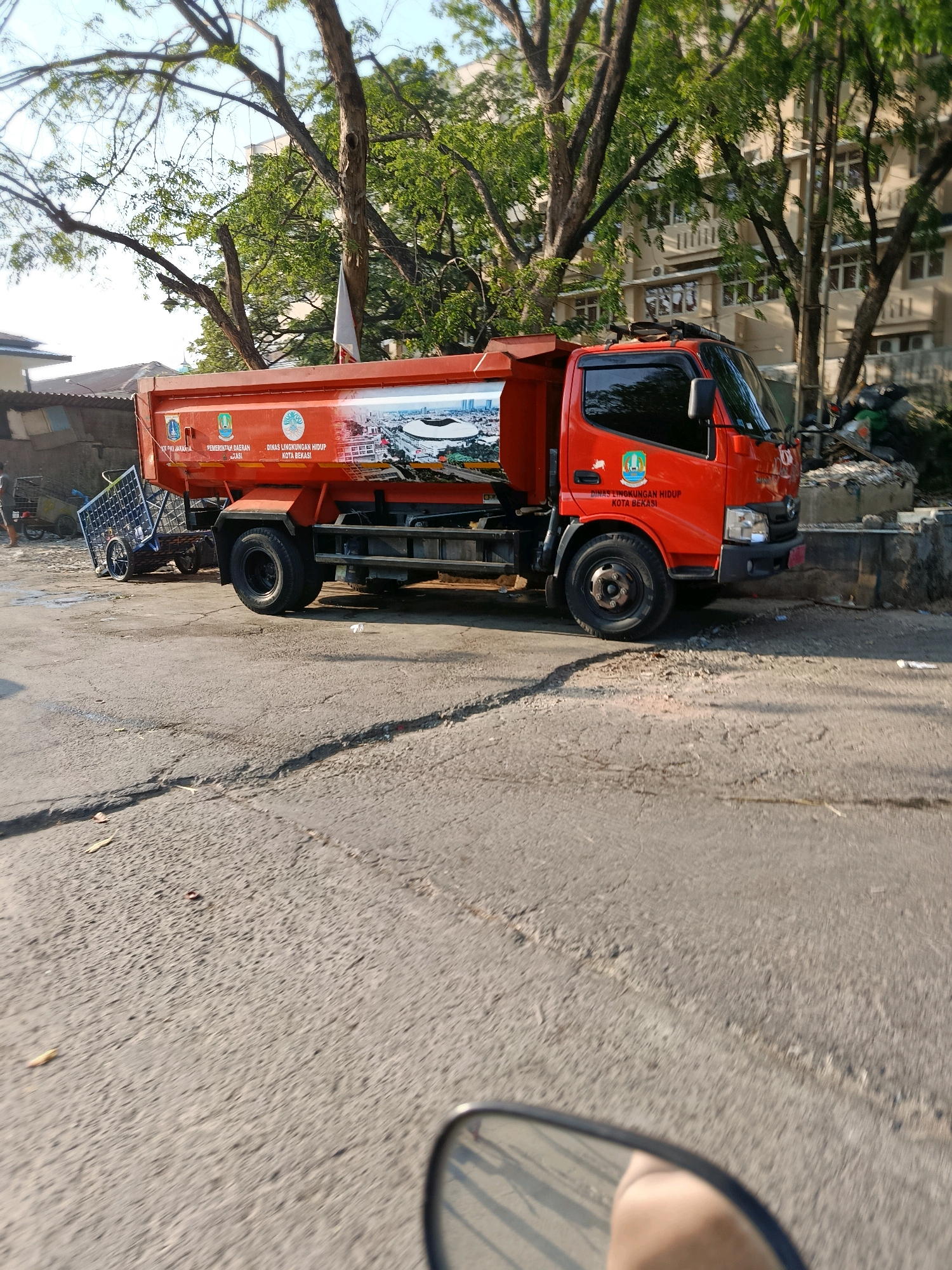 A bright orange garbage truck is parked on a cracked asphalt road in front of a building. The truck has graphics on its side depicting a city skyline and text that reads "PEMERINTAH DAERAH" and "DINAS LINGKUNGAN HIDUP KOTA BEKASI." A small white flag is attached to the top of the truck's cab. In the background, there are trees with green leaves and a multi-story building. To the left of the truck, a person is visible pushing a cart. The scene appears to be a street in the city of Bekasi, Indonesia, likely during daylight hours as indicated by the shadows.