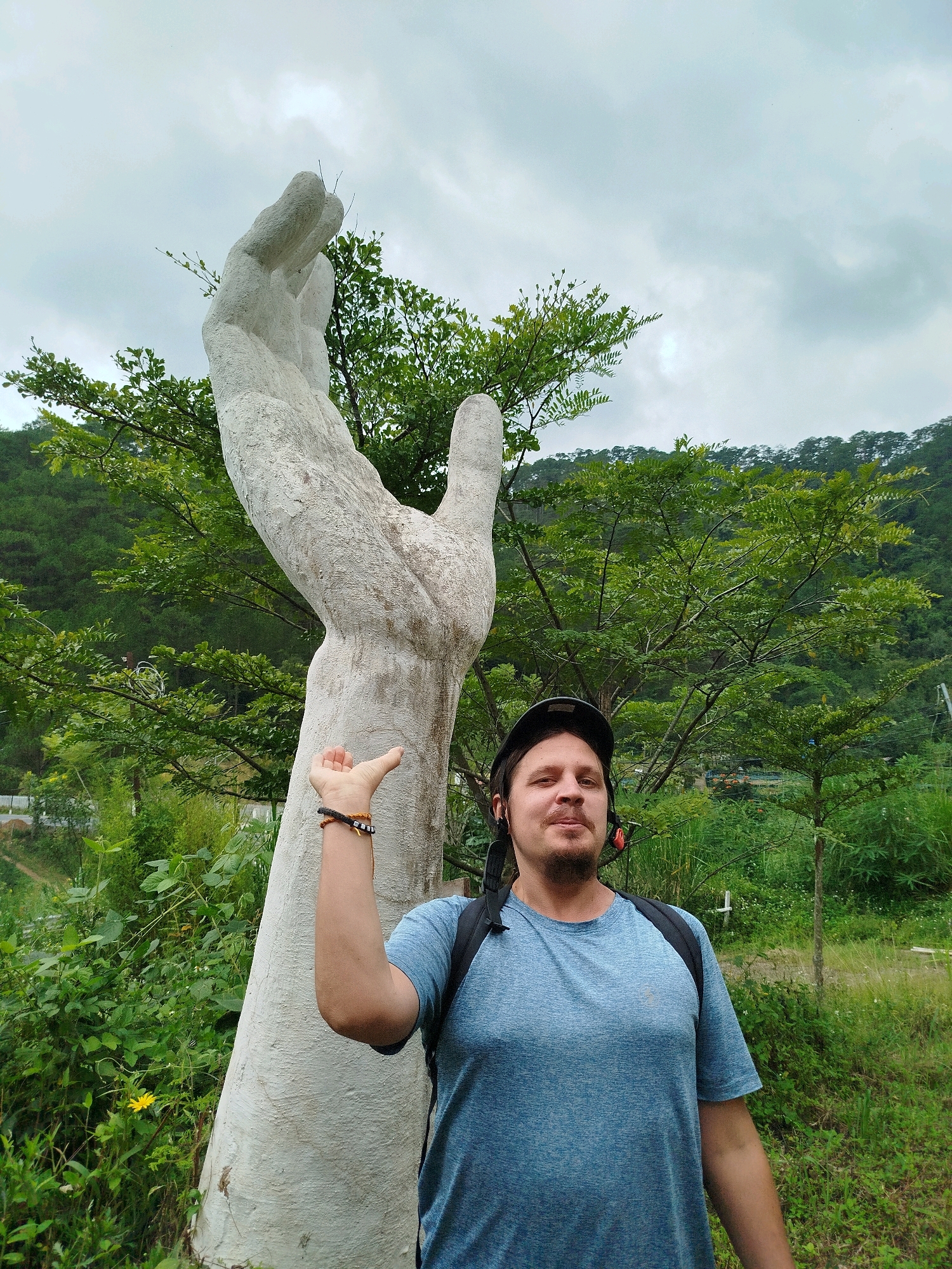 A man wearing a blue t-shirt and a black helmet poses in front of a large stone sculpture of a hand reaching upwards. The man is positioned to the right of the sculpture, with his left arm extended, thumb up, towards the hand. He is looking at the camera with a slight smile. The sculpture is textured and appears weathered. Behind them, a lush green landscape is visible, with trees, vegetation, and a hillside covered in trees under a cloudy sky. This scene is located at the hand sculpture at Tuyen Lam Lake in Da Lat, Vietnam.
