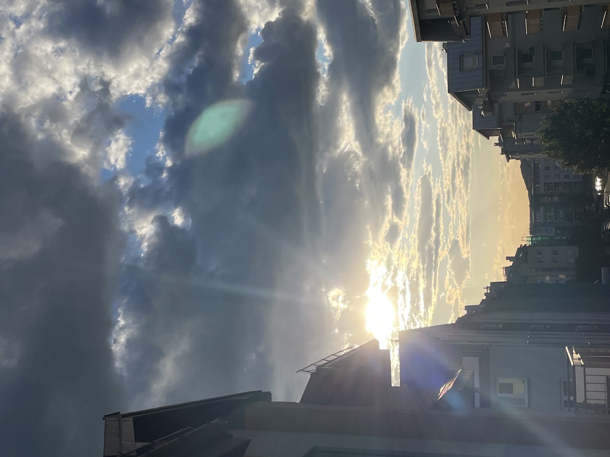 The image depicts a view upward from a street in an urban area. Tall buildings line the sides of the frame. The sky is filled with clouds, with the sun visible and casting rays of light through the clouds and onto the buildings. The location is Unknown.