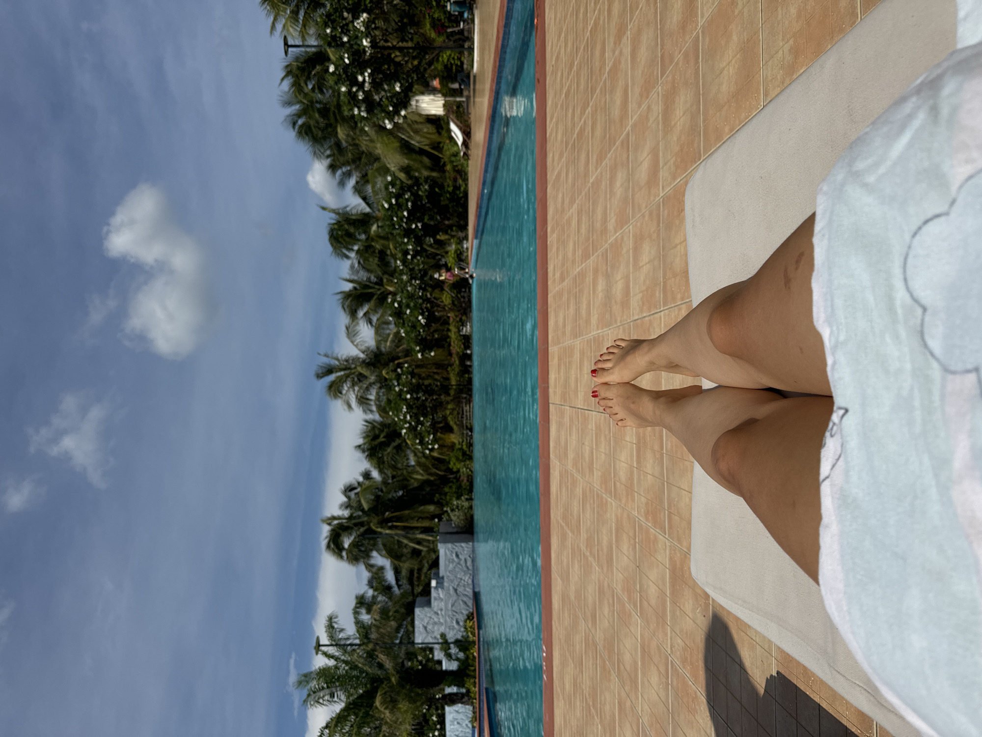 The image shows a first-person perspective of a person lying on a lounge chair by a swimming pool. The person's legs and feet are visible in the foreground, with red nail polish on the toenails. The swimming pool is turquoise blue and surrounded by a tiled patio. Palm trees and lush vegetation line the background, with a cloudy blue sky overhead. In the distance, there are some structures with a patterned facade and a few white lounge chairs. The scene appears to be a resort or a hotel in a tropical location, possibly Phu Thuy Ward, Vietnam, given the presence of palm trees and the overall ambiance. The lighting suggests it is daytime, with sunlight casting shadows on the patio.