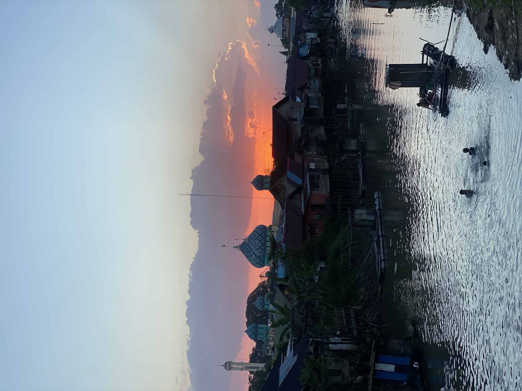 This photograph captures a sunset scene in Tunggul Irang Ulu, Indonesia. The sky is painted with hues of orange, pink, and pale blue, with fluffy clouds illuminated by the setting sun. In the foreground, a river flows, its surface rippling with the movement of water. Several individuals are visible in the river, some swimming and others gathered around a small raft. The raft appears to be constructed from logs and has a rudimentary structure on top, possibly a shelter or storage unit. A person is seen interacting with this structure on the raft.
Along the riverbanks, a settlement of houses is depicted. Many of these structures are built on stilts, indicating a potential flood-prone area. The architecture suggests a rustic and perhaps impoverished environment. In the background, several prominent buildings with domed roofs, characteristic of mosques, are visible. One mosque stands out with its distinctive blue domes and intricate patterns. A tall, slender minaret also punctuates the skyline. The overall impression is of a community living in close proximity to the river, with religious structures playing a significant role in the landscape. The time of day, indicated by the sunset, suggests it is late afternoon or early evening.