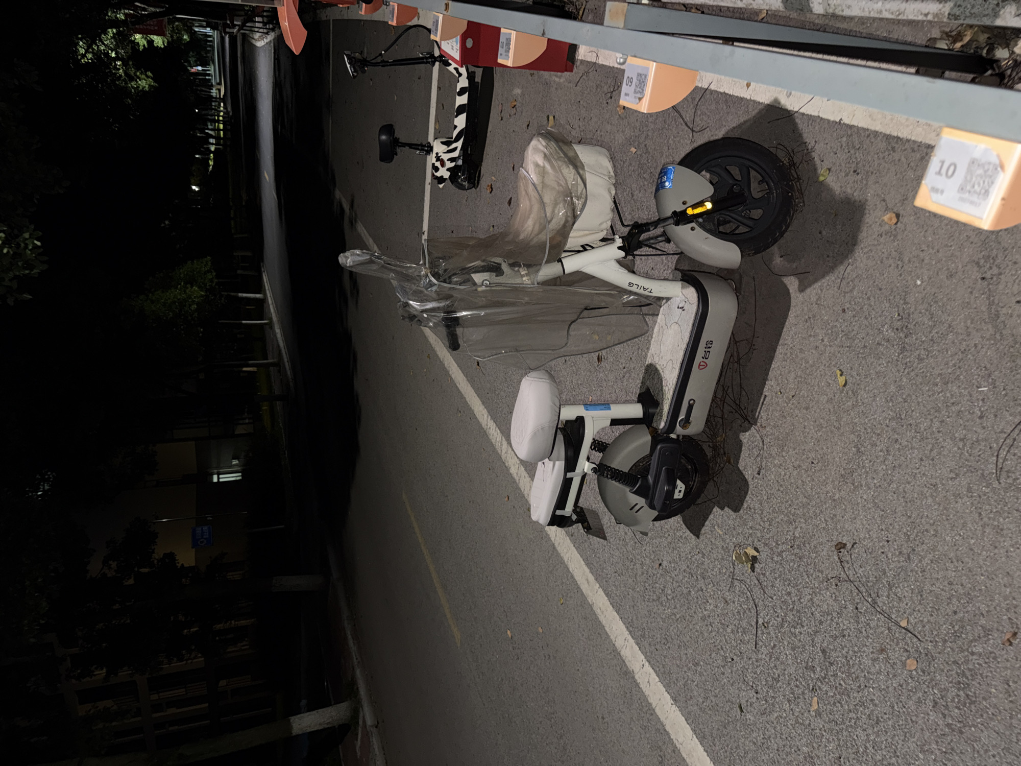 The image depicts an outdoor scene at night, likely in a public area within the city of Jiangjin, China, given the context of the provided information. The foreground is dominated by a white electric scooter parked on an asphalt surface, which appears to be a road or pathway. This scooter is partially covered by a clear plastic sheet. To the right of this scooter, another electric scooter with a black and white cow-print design is visible, parked in a designated charging or docking station. The docking station consists of orange colored units, some of which display QR codes. The background is dark, with trees lining the left side of the road and a building structure partially visible in the far distance. The lighting conditions suggest it is nighttime, with streetlights or ambient light casting shadows. There are no people present in the image.