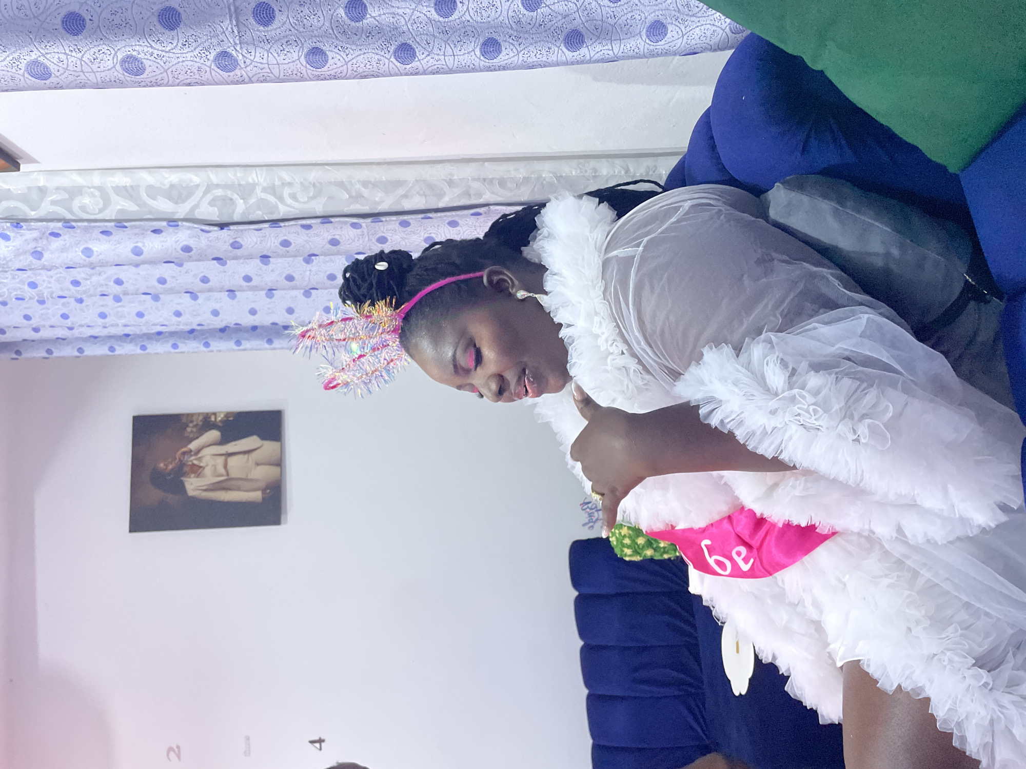 A woman wearing a white ruffled outfit and a pink crown sits on a blue couch. She has her hair in braids and is wearing pink eyeshadow. A pink sash that says "be" is draped over her outfit. She is holding her hand up as if to gesture. Behind her, there is a picture on the wall of a woman in a suit, and patterned curtains hang on the right side of the frame. A green cushion is visible to the right of the couch. The lighting in the room appears to be artificial.