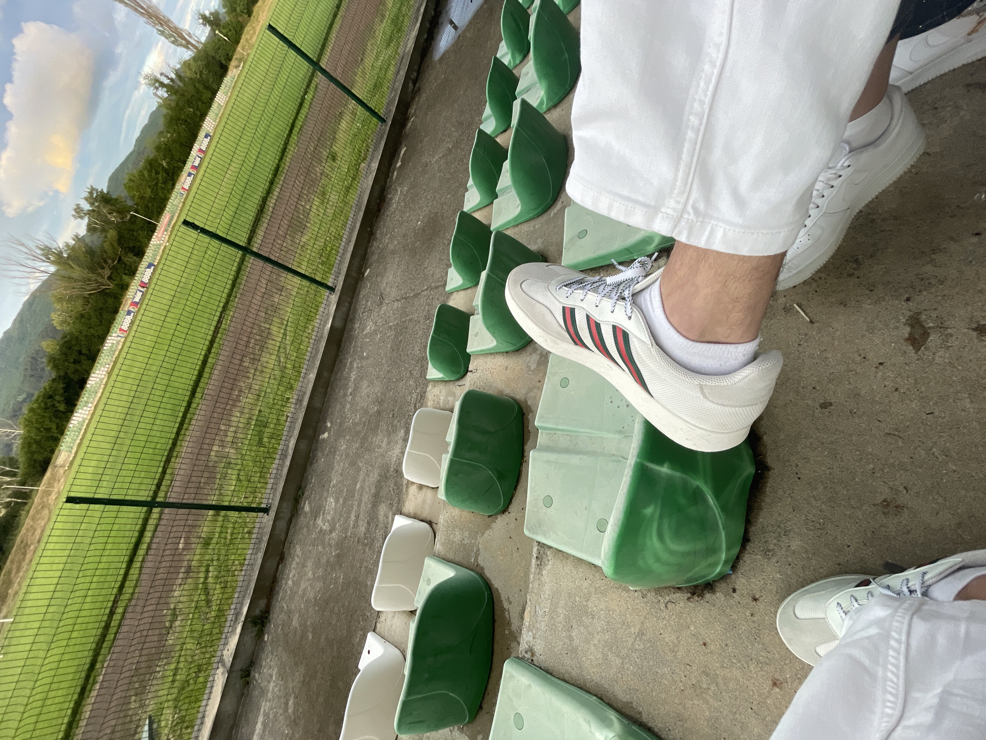 The image captures a partial view of a person in a sports stadium or arena. In the foreground, the lower legs and feet of a person are visible, wearing white loose-fitting pants, white socks, and white sneakers. One foot, adorned with a sneaker featuring prominent red and green stripes, rests on a green stadium seat. Another pair of white sneakers (appearing to be Nike Air Force 1s) is partially visible on the ground to the right.
The setting is an outdoor stadium stand, with rows of green and some lighter, possibly faded white, plastic seats. The seats are arranged on concrete steps, suggesting spectator seating. Beyond the stands, a green chain-link fence separates the spectator area from a sports field, likely a soccer or football pitch, with a dirt track visible alongside it. Along the far edge of the field, advertising boards are present, where the text "8888" is clearly visible in multiple repetitions. In the background, green trees and distant mountains rise under a blue sky, partially covered by a large white cloud. The lighting indicates it is daytime, likely sunny. No specific activity or event is taking place on the field, and no emotions are discernible from the partial view of the person.