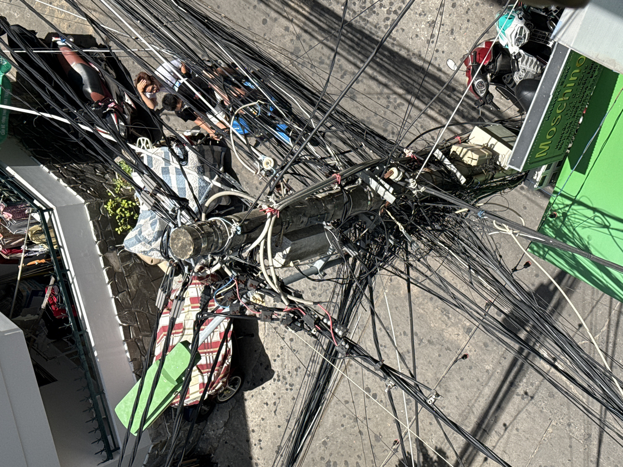 An overhead view captures a dense network of black electrical wires suspended above a street in Nha Trang, Vietnam. The wires are haphazardly strung between a central utility pole and various buildings. Below, on the concrete street, a red and black motorcycle is parked. Two individuals, one wearing a white shirt and the other a dark shirt, are standing near the motorcycle, appearing to interact with a third person partially obscured by the wires. Adjacent to the motorcycle, a green sign with yellow text displaying "Moschino" is visible. A green tarp-like object rests on what appears to be a cart or a low structure, partially covered by a striped fabric. The scene is bathed in sunlight, casting shadows from the wires onto the street.