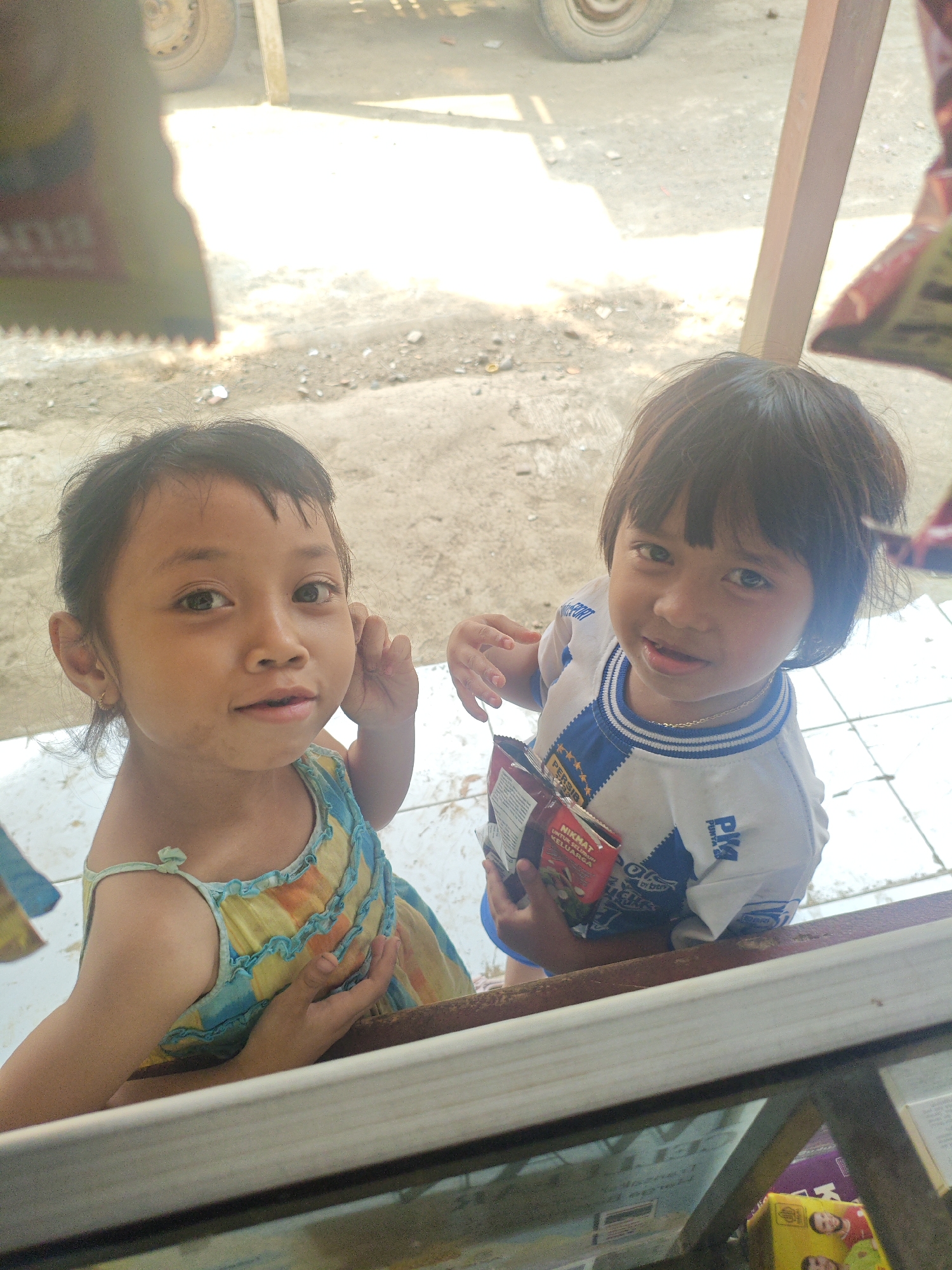 Two young girls are standing in front of a display case, likely at a small shop in Bantarwaru, Indonesia. The girl on the left, with dark hair and a small gold earring, wears a colorful yellow, blue, and orange dress. She is looking directly at the camera with a slight smile. The girl on the right, with dark hair and bangs, is wearing a white shirt with blue trim and lettering. She is holding a small, red and white package in her hands and looking towards the camera. The background appears to be an outdoor area with a dirt or gravel ground and a wooden post. The lighting suggests it is daytime, possibly late morning or early afternoon, with strong sunlight creating some bright areas and shadows. The overall scene is casual and appears to be a candid photograph.
