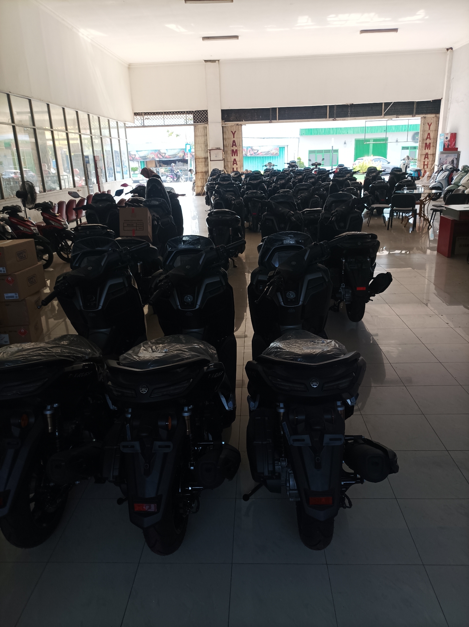 The image depicts the interior of a Yamaha motorcycle dealership in Ploso, Indonesia. Rows of new, black Yamaha motorcycles are parked on a tiled floor, with some covered in plastic. Boxes are stacked to the left of the motorcycles, some bearing "SAFETY TOOLS" labels. In the background, a person in a headscarf is visible near the entrance, and several other motorcycles are parked outside. The dealership appears to be a spacious, well-lit area with large windows and metal grilles. The overall impression is of a busy retail environment showcasing a large inventory of motorcycles.