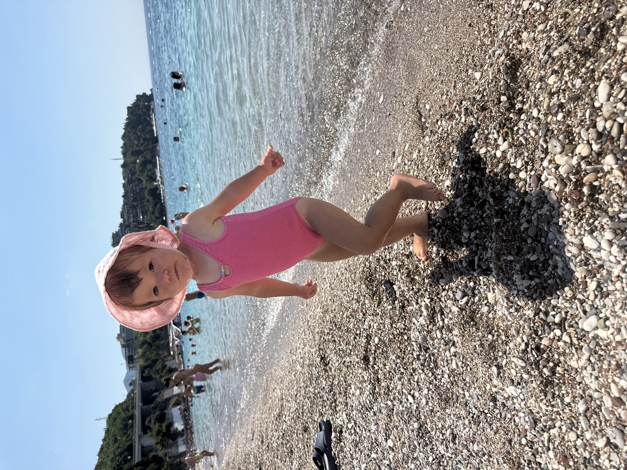 In this eye-level, outdoor, vertical-orientation photograph, a young child is the primary subject, standing on a pebble beach next to the sea. The child, likely a toddler, is wearing a pink one-piece swimsuit and a matching pink sun hat. Their gaze is directed towards the camera with a neutral expression. The child's left arm is bent and raised, with their hand in a fist, while their right leg is extended behind them, suggesting a playful or dynamic pose.
The setting is a beach with a clear, shallow sea. The water exhibits gentle ripples and reflections from the sunlight. The shoreline is composed of numerous small, smooth pebbles in varying shades of grey, white, and brown. In the background, beyond the water, a lush, green hillside with trees is visible, dotted with buildings and a bridge. Several other people are present in the water and on the shore, some appearing to be swimming or wading.
The bright sunlight suggests it is daytime, likely midday or afternoon, given the quality of light and the clear blue sky. The scene evokes a summery, vacation atmosphere. The location is identified as Boreti, Montenegro, which is consistent with the scenic coastal environment depicted. No visible text is present in the image.