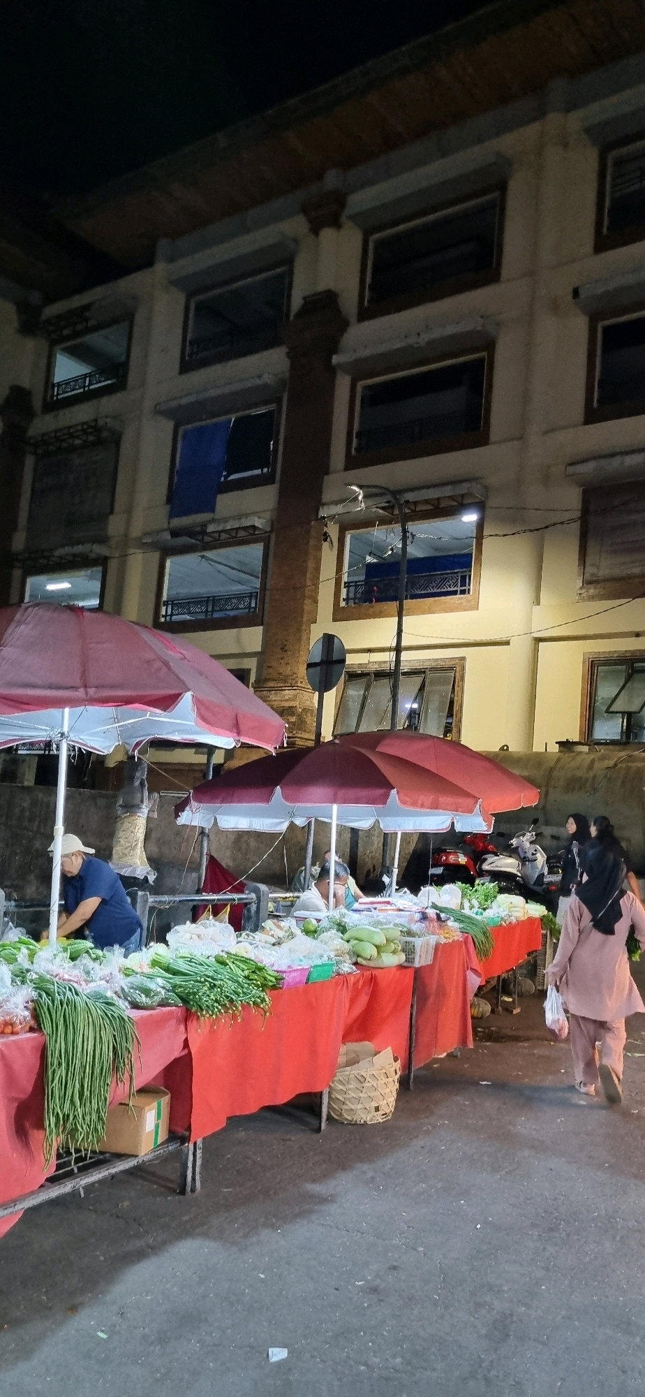 This image depicts an outdoor market scene at night, likely in Denpasar, Indonesia, as evidenced by the architecture and overall atmosphere. The focal point is a row of market stalls displaying an abundance of fresh vegetables such as long beans, onions, and squash. Two large red and white umbrellas provide shade over the stalls.
A man in a blue shirt and white cap is visible behind one of the stalls, appearing to be tending to the produce. Further down the line, a woman in a light brown headscarf and matching outfit is walking away from the camera, carrying a plastic bag. Behind her, another woman in a dark headscarf and dark clothing is also walking. In the background, a multi-story building with numerous windows is illuminated, suggesting it is an urban setting. The street is paved, and shadows from the stalls and umbrellas are cast on the ground. The lighting appears to be a mix of ambient light from the building and possibly stall lighting, creating a slightly dim but functional environment for nighttime commerce. Several motorcycles are parked in the background, indicating a typical mode of transportation in the area.