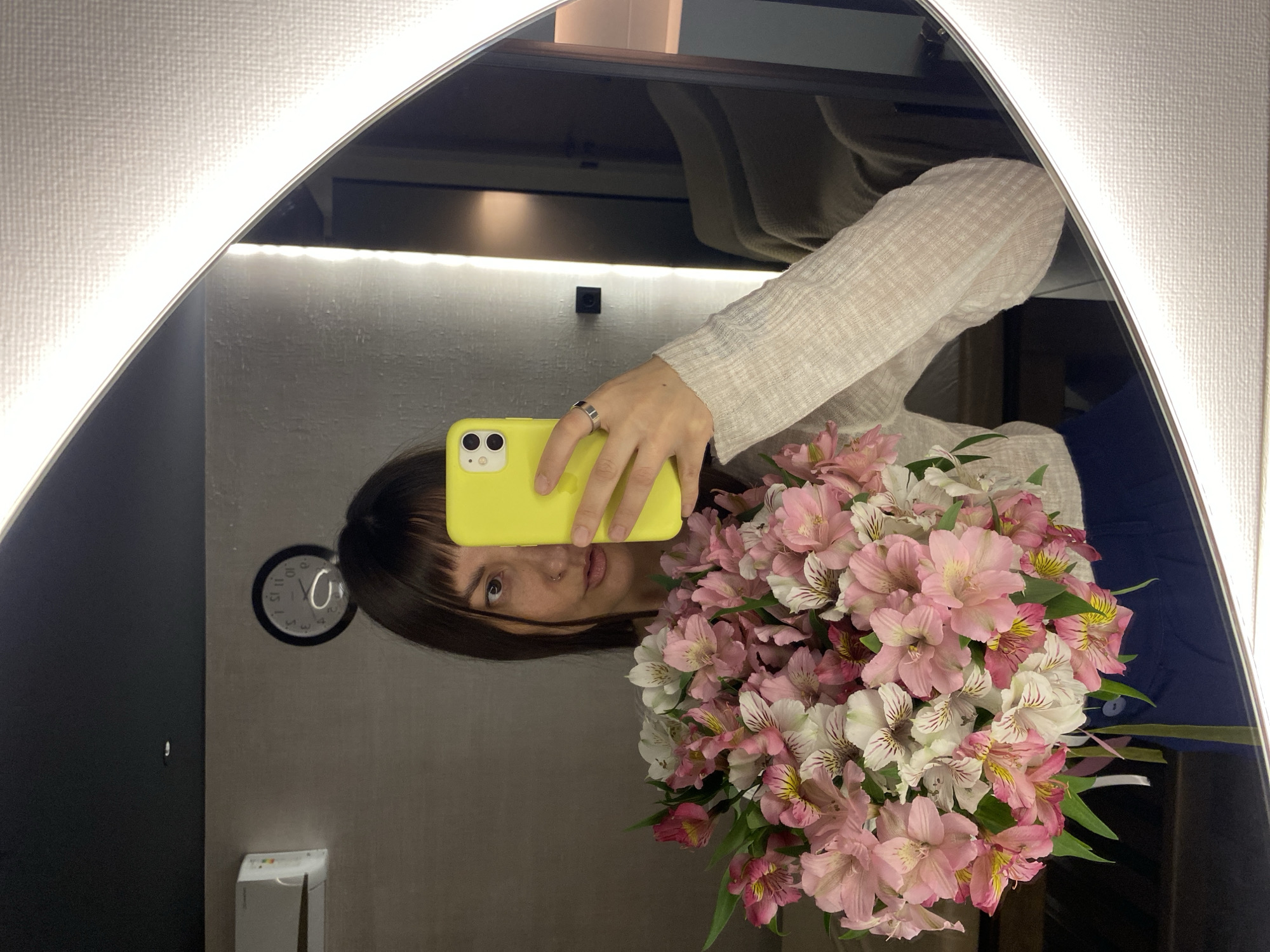 A person is taking a selfie in a mirror, holding a bouquet of pink and white alstroemeria flowers in front of their face. The person is wearing a light-colored, long-sleeved shirt and a blue garment is visible behind the flowers. The mirror is surrounded by a white, illuminated frame. A clock is visible on the wall behind the person, and there is a hand sanitizer dispenser below the clock. The environment appears to be an indoor space, possibly a fitting room or a bathroom. It is not possible to determine if the location is in Votkinsk, Russia, based on the given image.