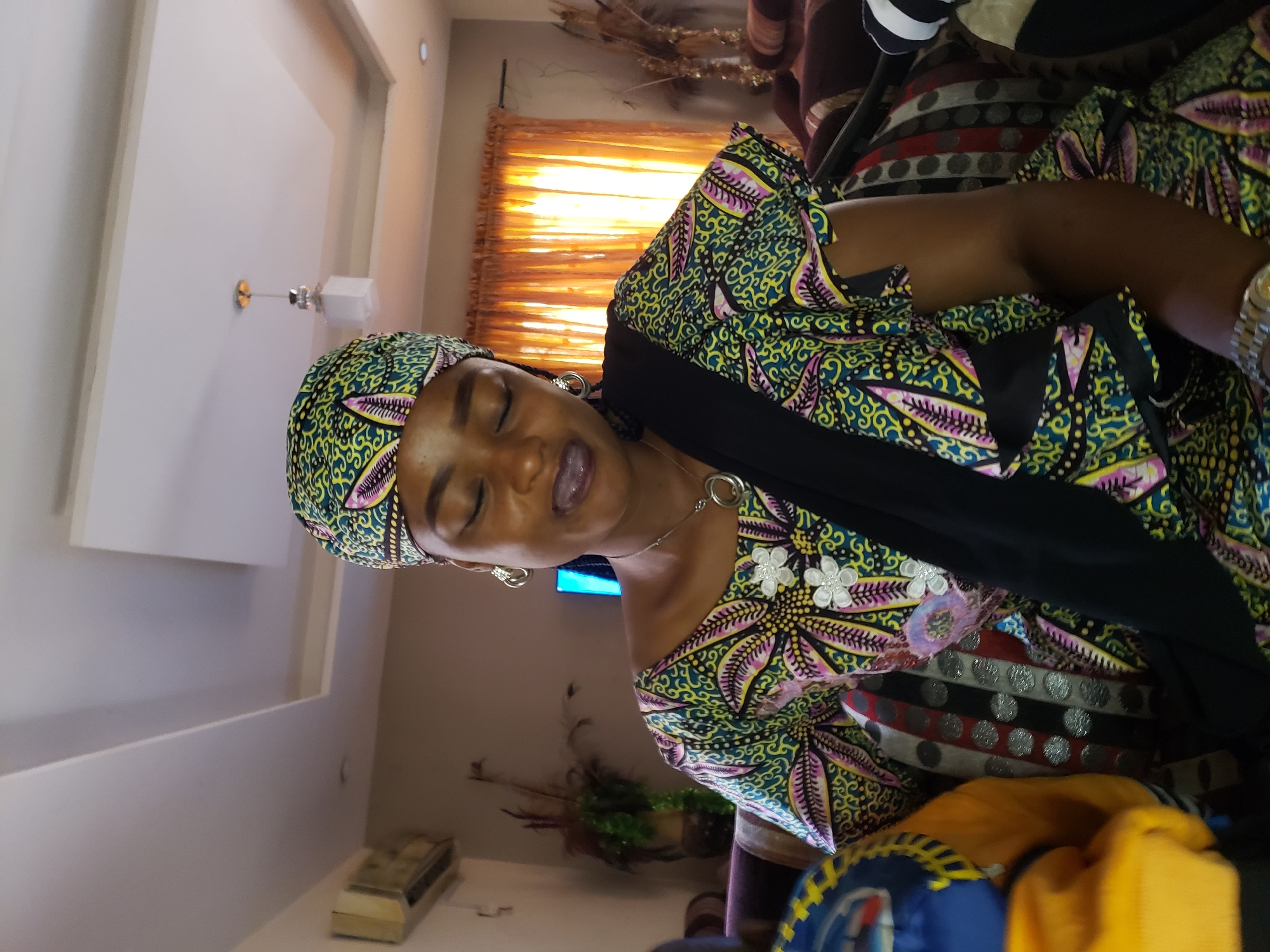 A medium shot captures a young woman from the shoulders up, seated indoors. She wears a vibrant, multi-colored headscarf and matching dress, both adorned with intricate floral and geometric patterns in green, yellow, pink, and black. Her eyes are closed, and her tongue is slightly extended, suggesting a moment of playful contemplation or perhaps a reaction to something she is tasting. She has earrings and a delicate silver necklace with a circular pendant. A black sash is draped across her chest, over her patterned dress.
The background reveals a residential interior. To the left, an air conditioning unit is visible on a beige wall. Further back, tall, thin dried plants are arranged in a vase, creating a naturalistic element. The ceiling features recessed lighting and a modern, square light fixture with a golden base. Orange curtains are drawn across a window, allowing soft, diffused light to enter the room. The woman is seated on a patterned sofa, with other pillows and furniture visible in the dimly lit background. The overall scene suggests a casual setting, possibly within a home in the Sabon Tasha area of Nigeria.