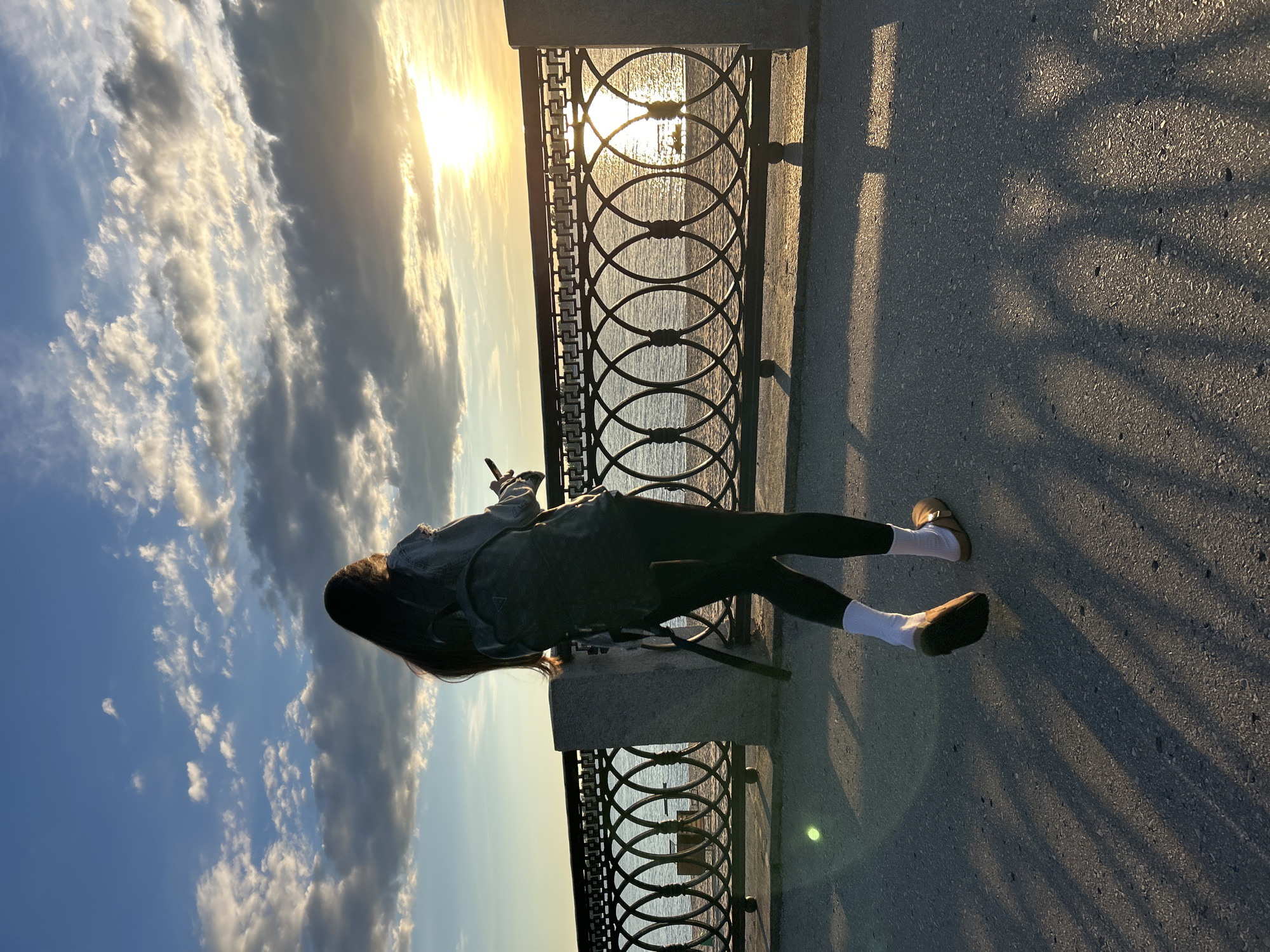 The image depicts a young woman with long dark hair, viewed from behind and from a high angle, as she is lying on a concrete surface next to a metal fence overlooking a body of water. She is wearing a denim jacket, black leggings, and white socks with brown shoes. The sun is setting, casting long shadows on the ground and illuminating the sky with warm golden light. The clouds are fluffy and white, suggesting fair weather. The fence has a decorative pattern of interlocking circles. The scene appears to be a waterfront promenade or embankment. The overall mood is peaceful and serene, possibly capturing a moment of quiet contemplation or relaxation by the young woman. There is no visible text in the image.