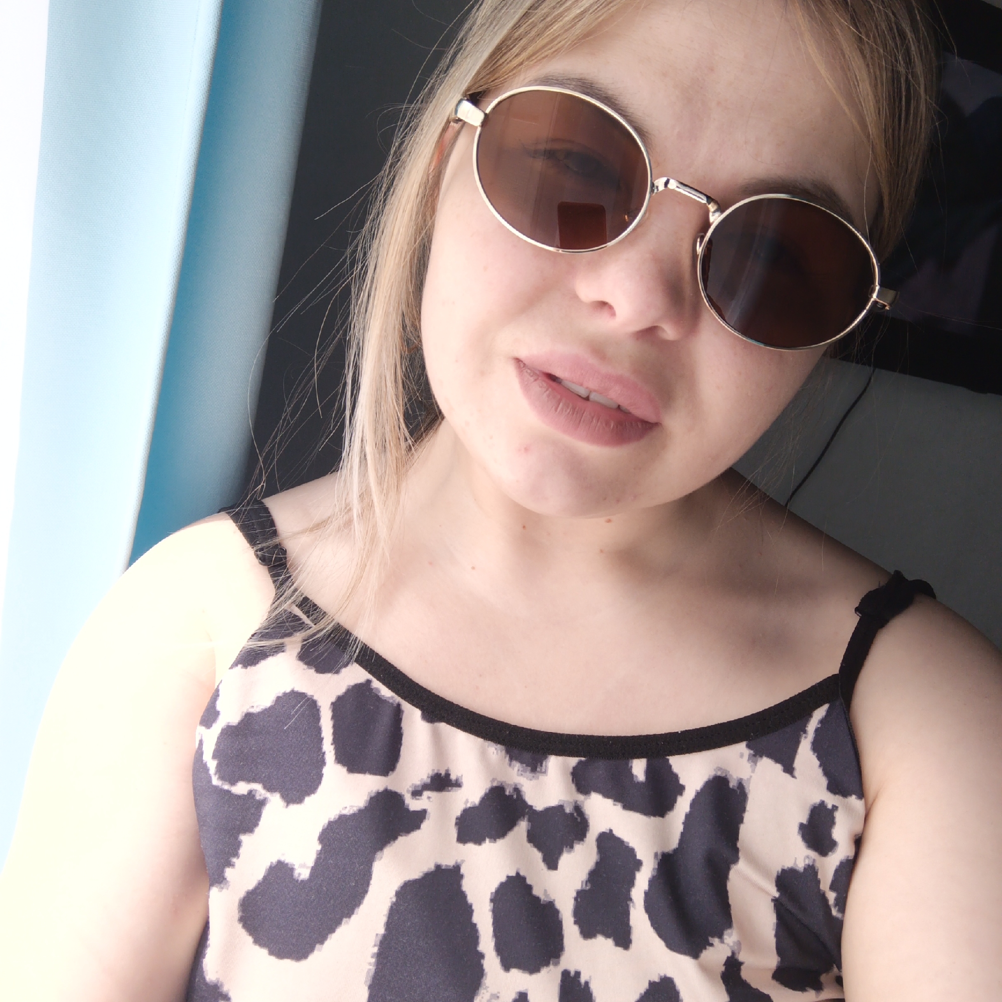 A close-up, slightly angled portrait features a young woman wearing round, gold-rimmed sunglasses with brown tinted lenses. Her blonde or light brown hair is visible around her face. She has light pink lipstick on her lips, which are slightly parted. Her skin shows some minor blemishes.
She is wearing a camisole or top with a distinctive animal print pattern, characterized by irregular black or dark gray spots on a light beige or off-white background, possibly resembling cow print. The top has a black trim around the neckline and thin black spaghetti straps.
The background on the left side of the image is dominated by a light blue, textured fabric, likely a curtain or blinds, suggesting she is indoors near a window. The right side of the background is darker and less defined, appearing as an indistinct indoor area. The lighting suggests it is daytime. No visible text is present in the image.