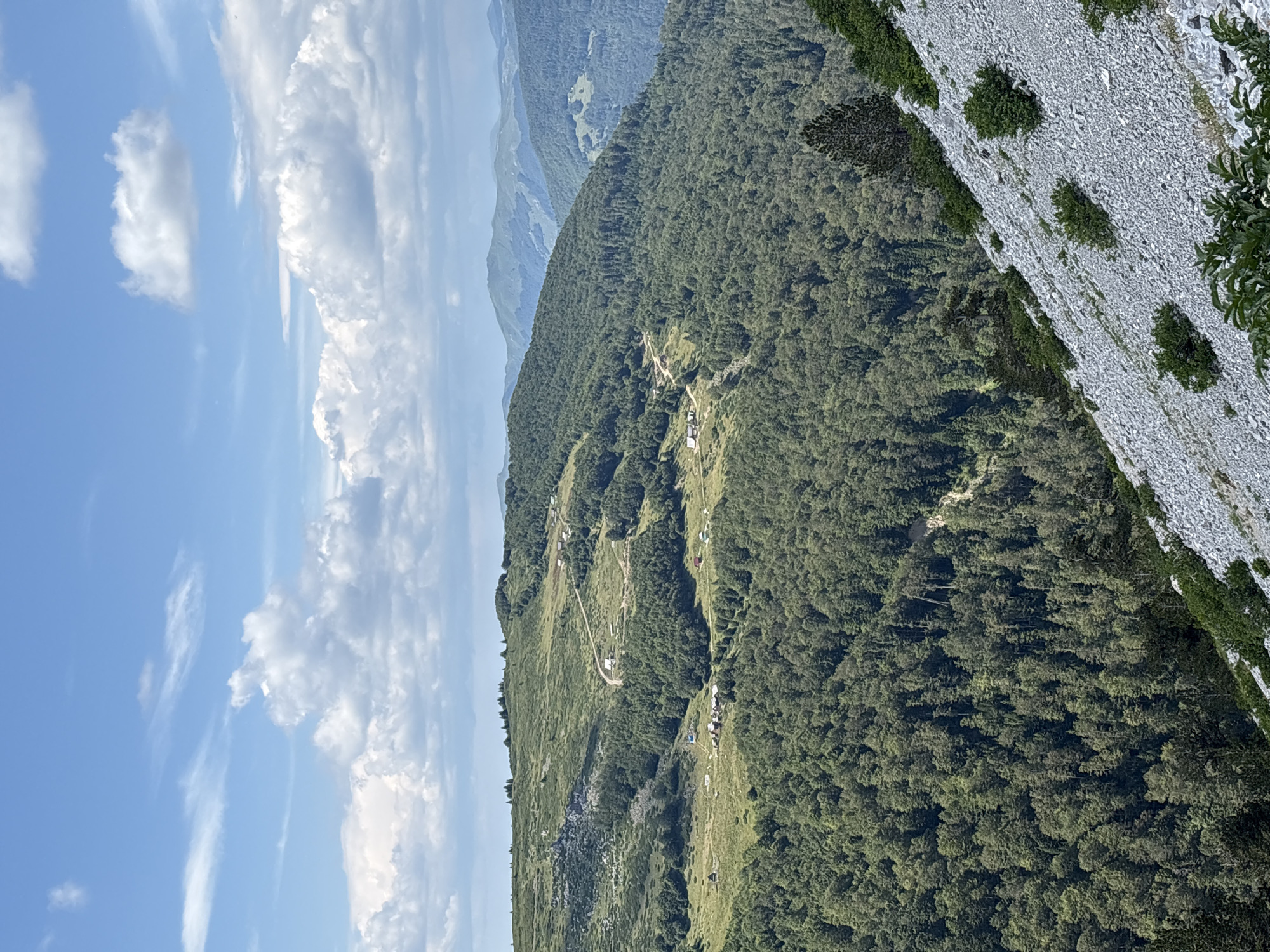 The image depicts a panoramic view of a mountainous landscape near Katun Vulića, Montenegro. The foreground is dominated by a steep, rocky slope covered in small grey stones and sparse green vegetation. In the middle ground, a densely forested hillside rises, with a winding dirt road cutting through the trees and leading to a cluster of small buildings, possibly houses or shelters. Further into the distance, rolling green hills are visible, covered in more forests and grasslands, with a hazy mountain range forming the backdrop under a partly cloudy blue sky. The scene conveys a sense of remote wilderness and natural beauty.