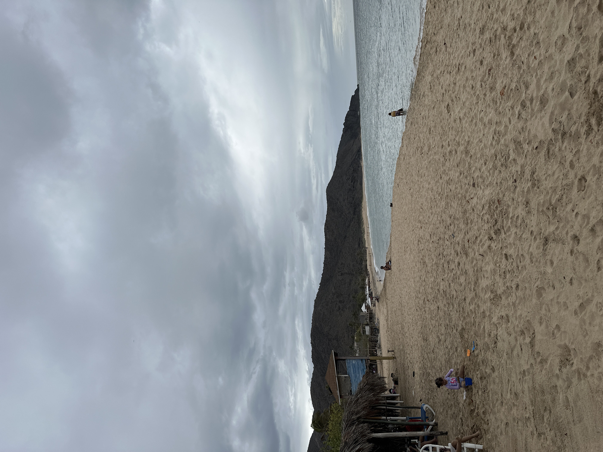 The image depicts a cloudy day at a beach located in Altagracia, Venezuela. The foreground is dominated by a sandy beach with footprints and scattered debris. In the mid-ground, a young girl is playing in the sand with toys. To the left, several thatched-roof structures provide shade, with some beach chairs visible. Further along the beach, in the distance, a few people are scattered, some near the water's edge and others sitting on the sand. The water is calm, with small waves lapping the shore. The background features a long, undulating mountain range covered in vegetation, sloping down to the coastline. The sea stretches to the horizon, meeting a sky filled with heavy, gray clouds, suggesting an overcast or impending rainy weather.